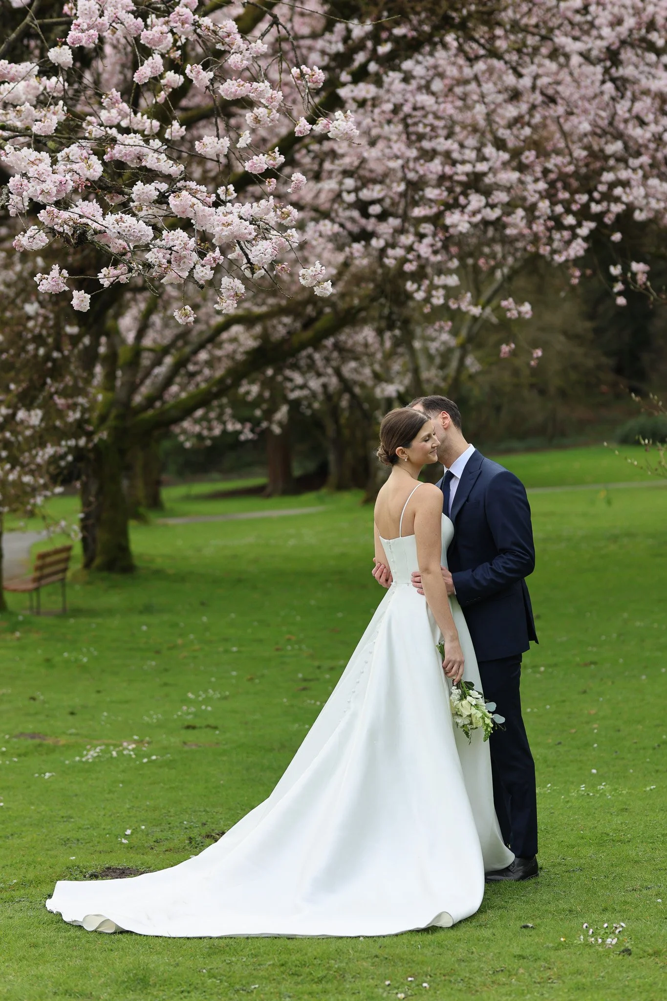 A bride and groom stand close together in a park with lush green grass and trees blooming with pink and white cherry blossoms. The bride, in a white wedding gown and holding a bouquet, leans into the groom, who is in a navy blue suit, as they share a