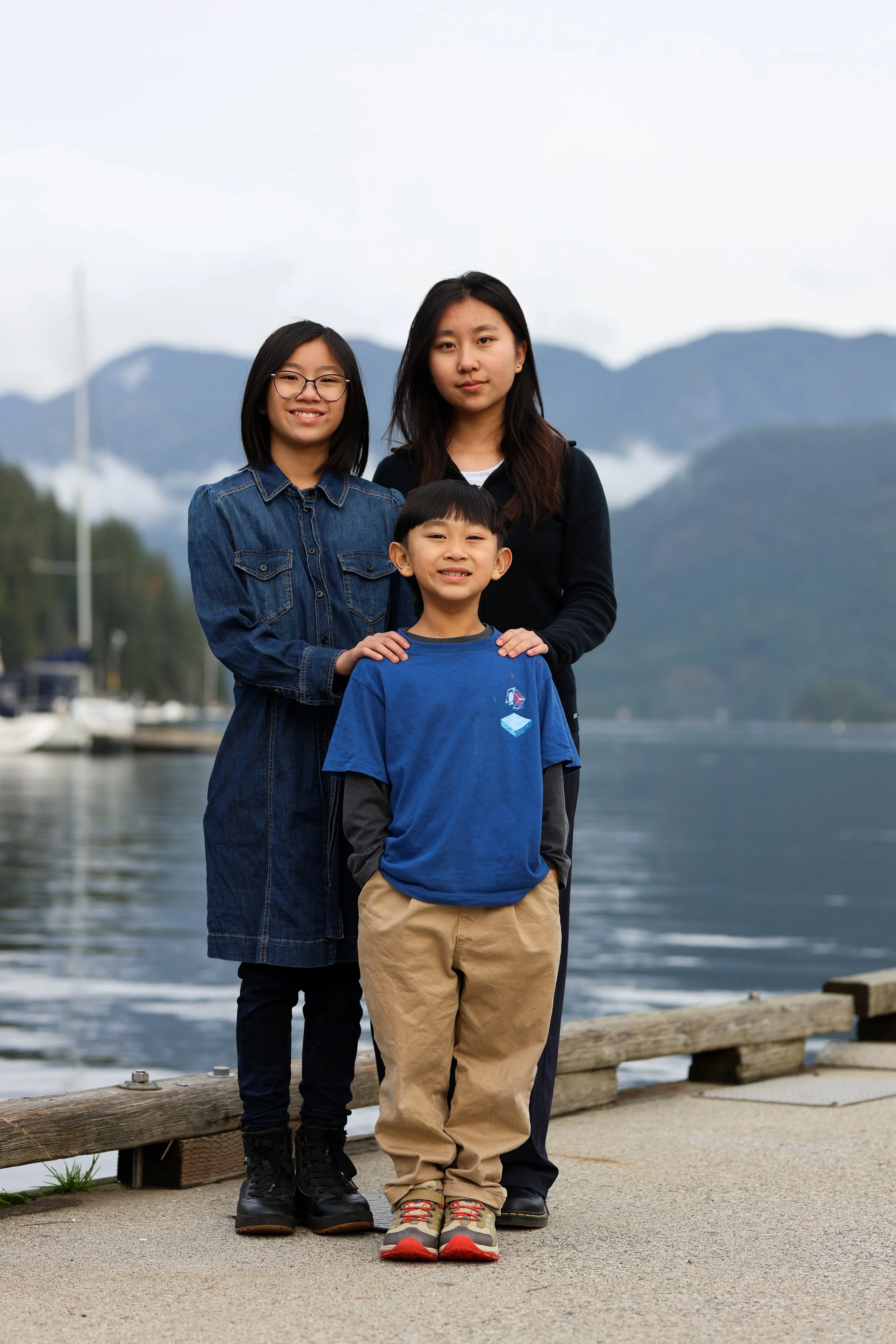 Three children standing outdoors on a paved area near water and mountains. The girl on the left is wearing glasses, a denim dress, and black boots. The boy in front is wearing a blue t-shirt, khaki pants, and sneakers. The girl on the right is wearin