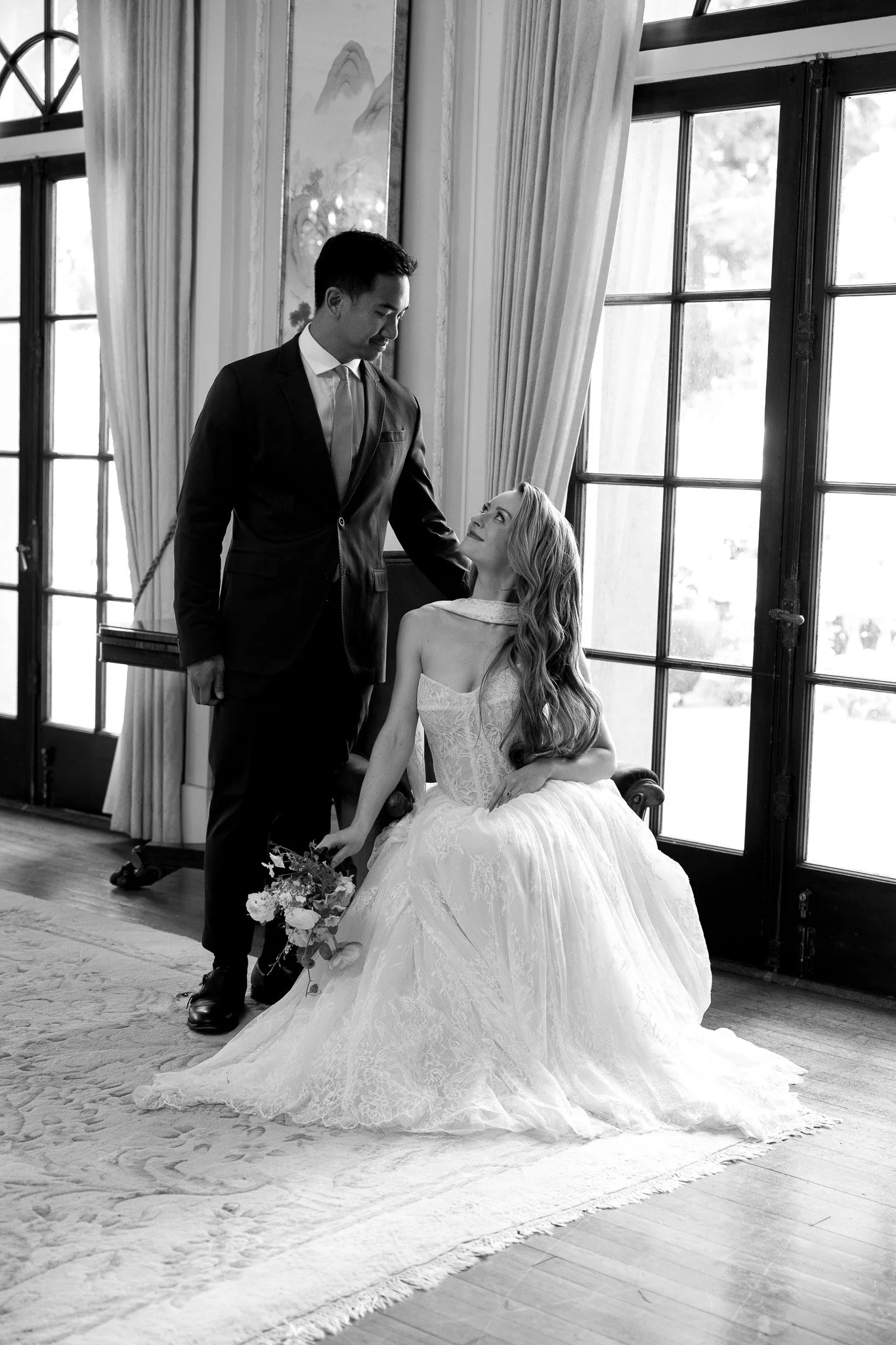 A black and white photo of a bride sitting on a chair, wearing a white lace wedding gown and holding a bouquet of flowers, looking up at a groom standing beside her, inside a room with large windows and curtains.
