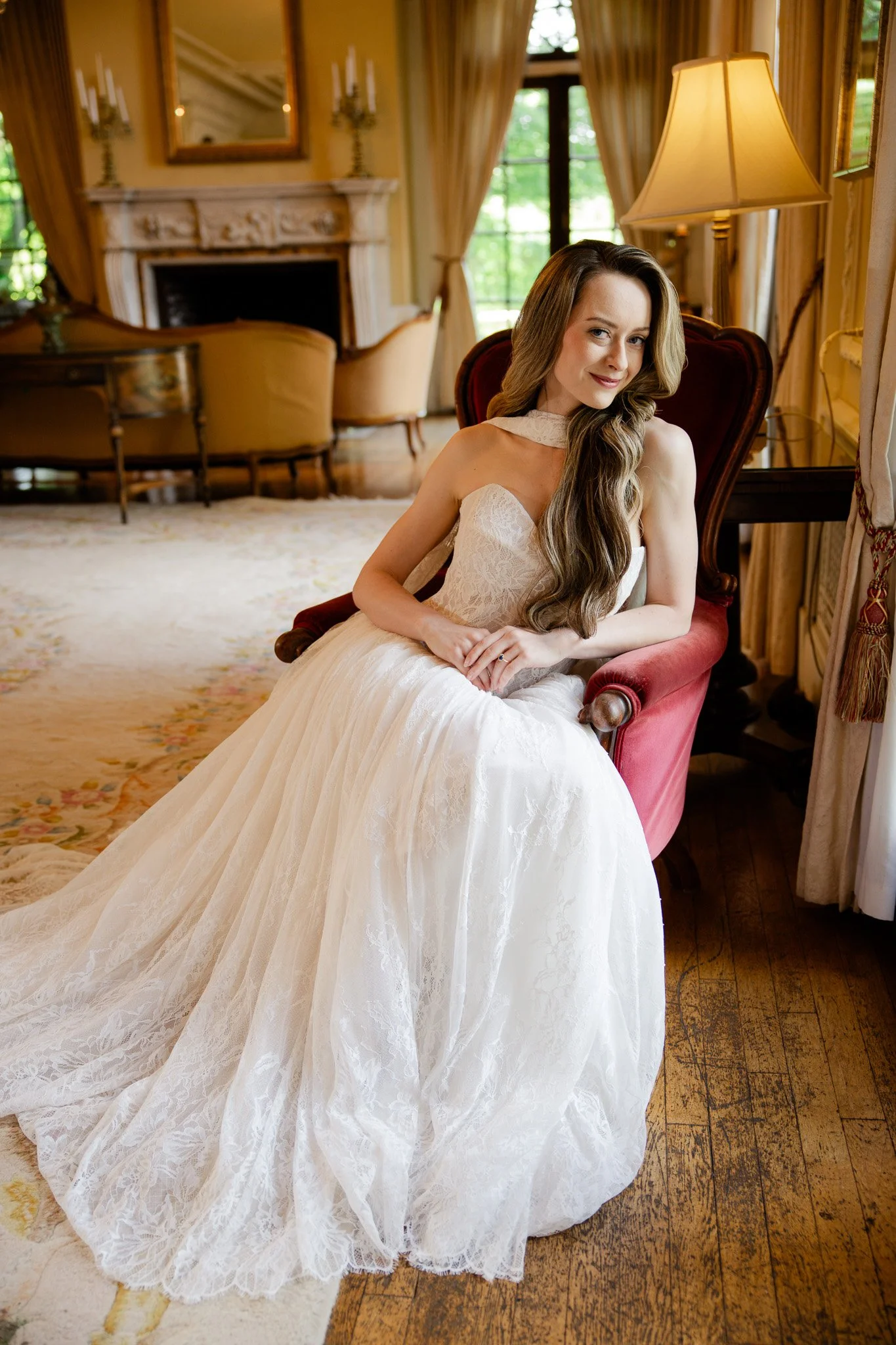A woman in a white wedding dress sitting in a vintage-style room with yellow and beige decor, a fireplace, and a large window with curtains, smiling at the camera.