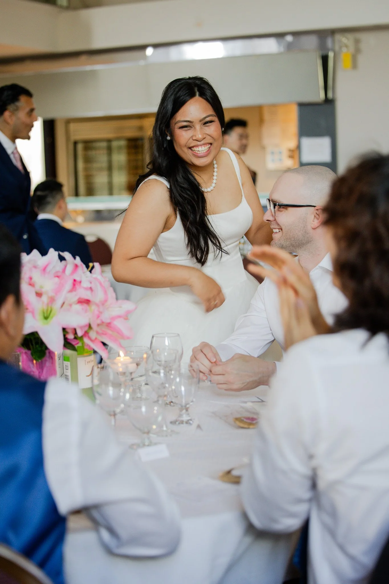 A woman in a white dress and pearl necklace smiling and talking to a person wearing glasses at a wedding reception table, with pink flowers and candles on the table.