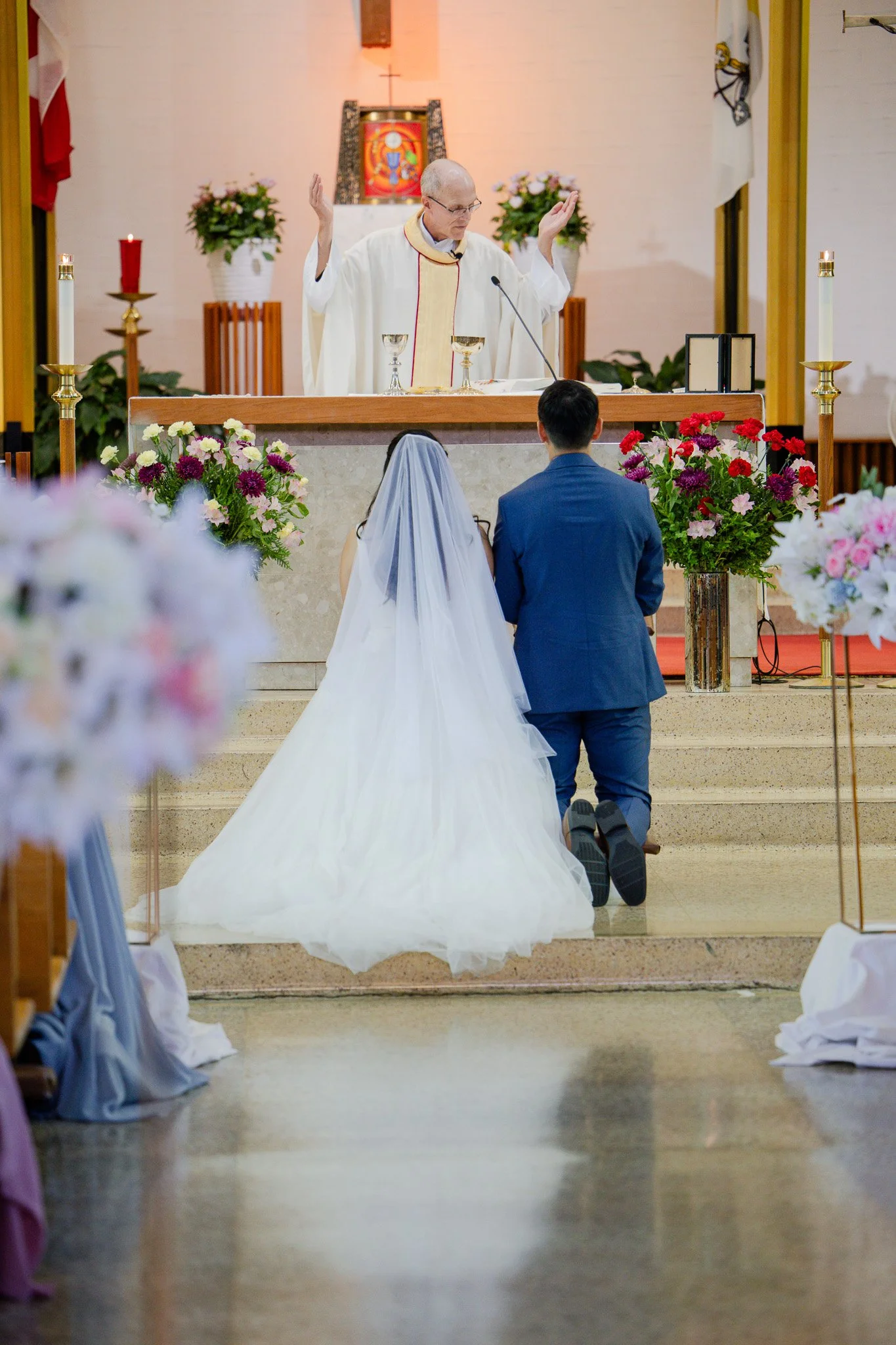 A wedding ceremony taking place in a church with a priest conducting the vows. The bride, dressed in a white gown with a long train and veil, and the groom, in a blue suit, kneel facing the altar decorated with flower arrangements.