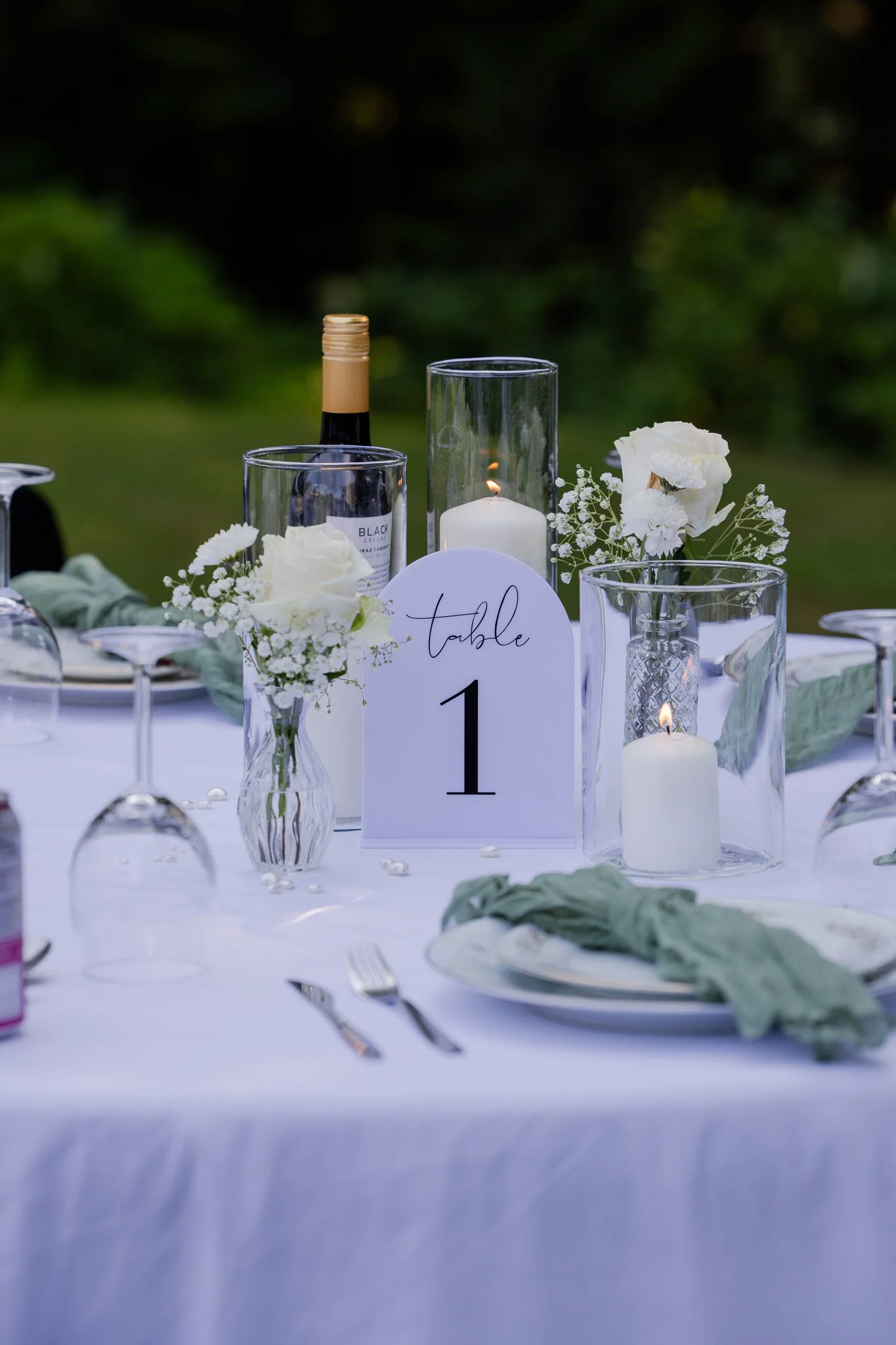 Elegant outdoor table setting with white flowers, candles, wine bottle, and numbered table card on a white tablecloth.