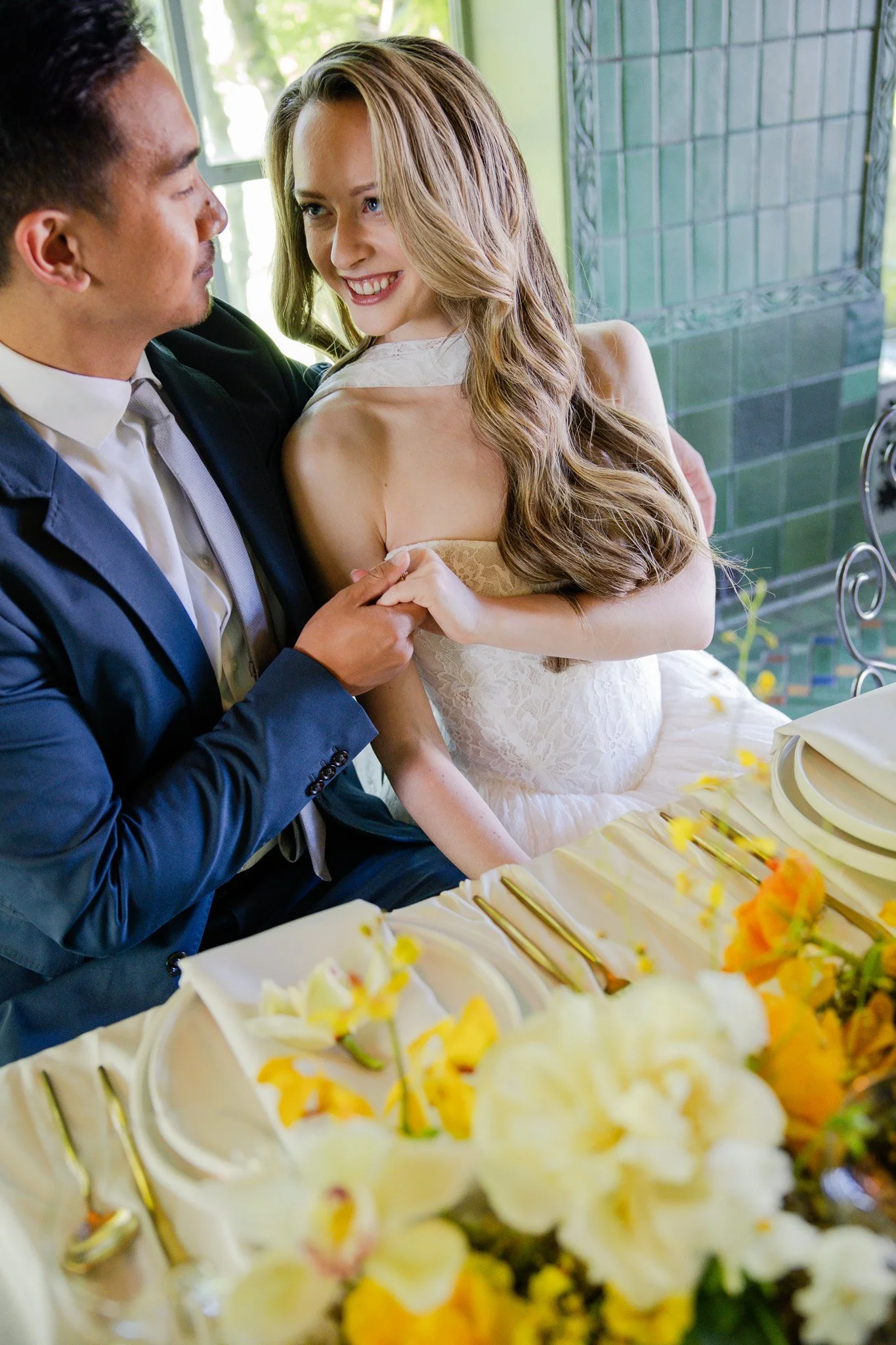 A man and woman share a joyful moment at a wedding reception, with the man dressed in a suit and the woman in a wedding dress. They are in close proximity, smiling, and holding hands, with a decorated table featuring cake and flowers in the foregroun