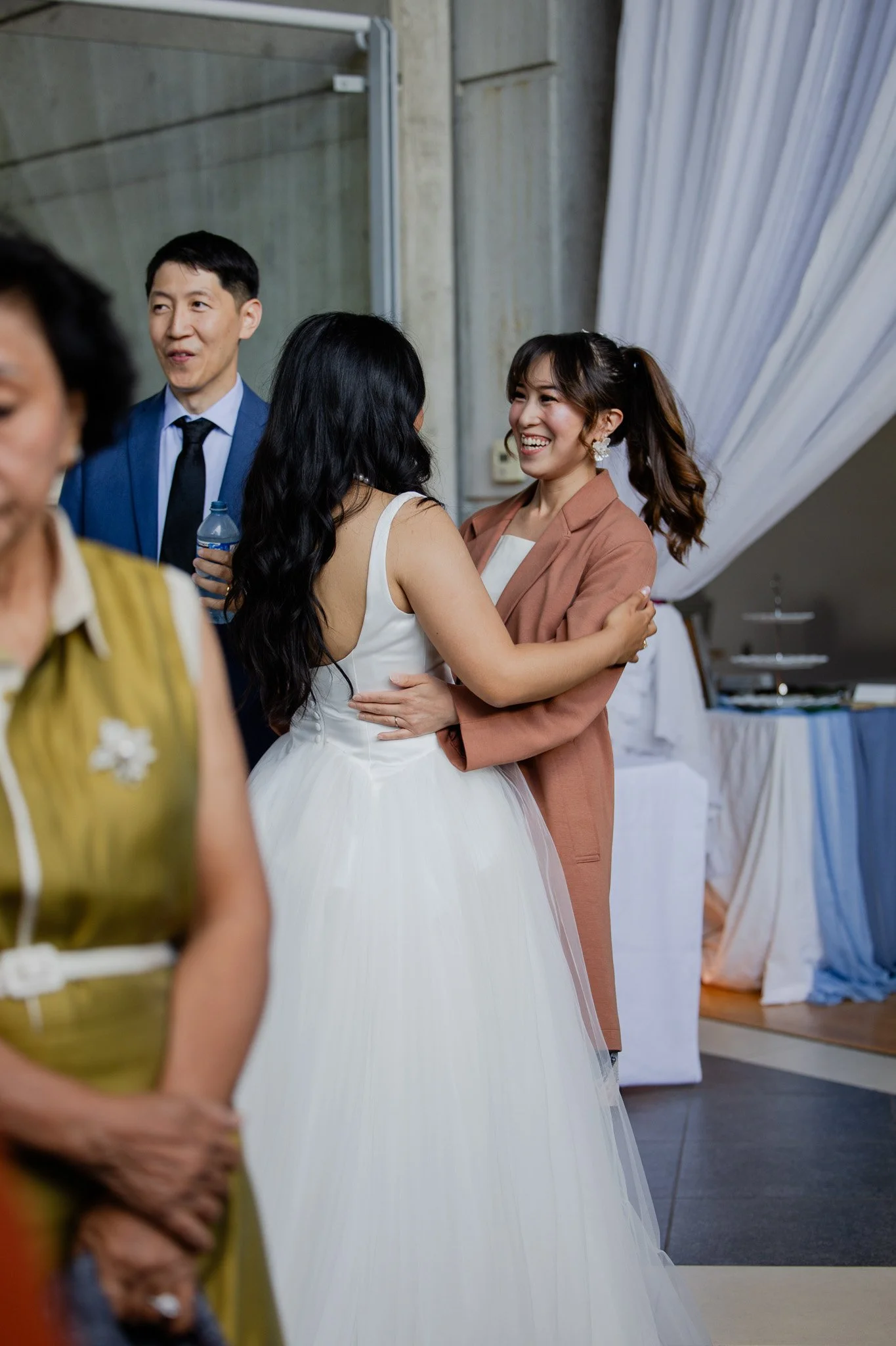 Two women and two men at a social gathering indoors, smiling and chatting. One woman wears a white dress, and the other woman wears a brown blazer. One man wears a blue suit with a black tie, and the other person's face is partially out of the frame,