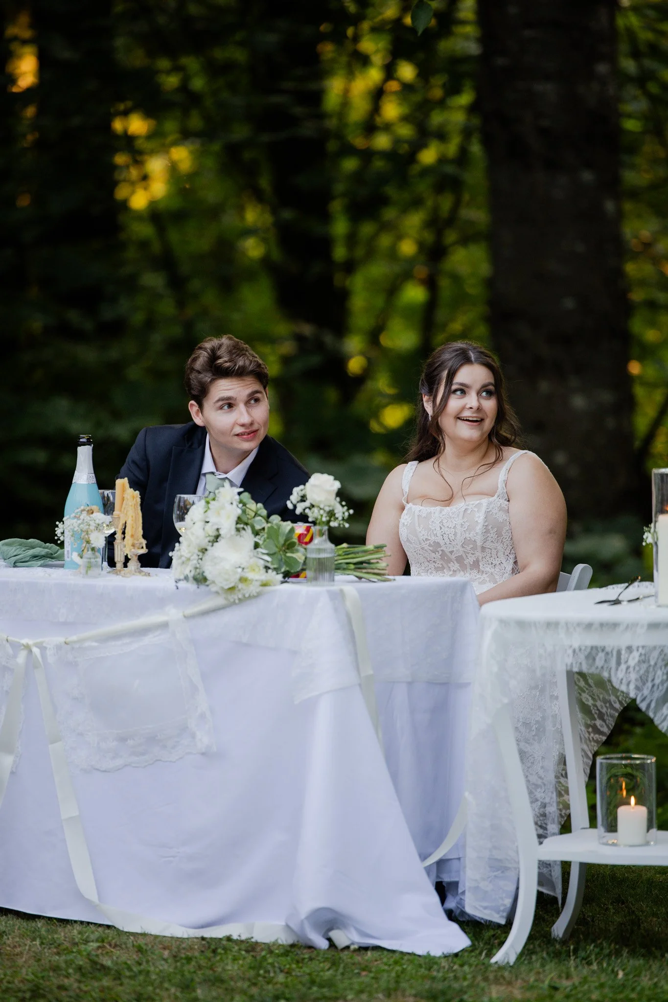 A man and woman are seated at a white table outdoors in a wooded area. The man wears a dark suit and white shirt, and the woman wears a white lace dress. The table is decorated with white flowers, candles, and various drinks, and the setting appears 