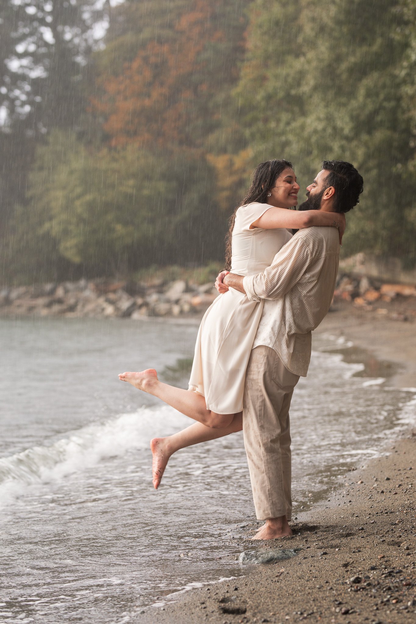 A couple is standing in the shallow water at the beach, smiling and embracing as the man lifts the woman off the ground. It is raining softly, and the background features trees with autumn foliage.