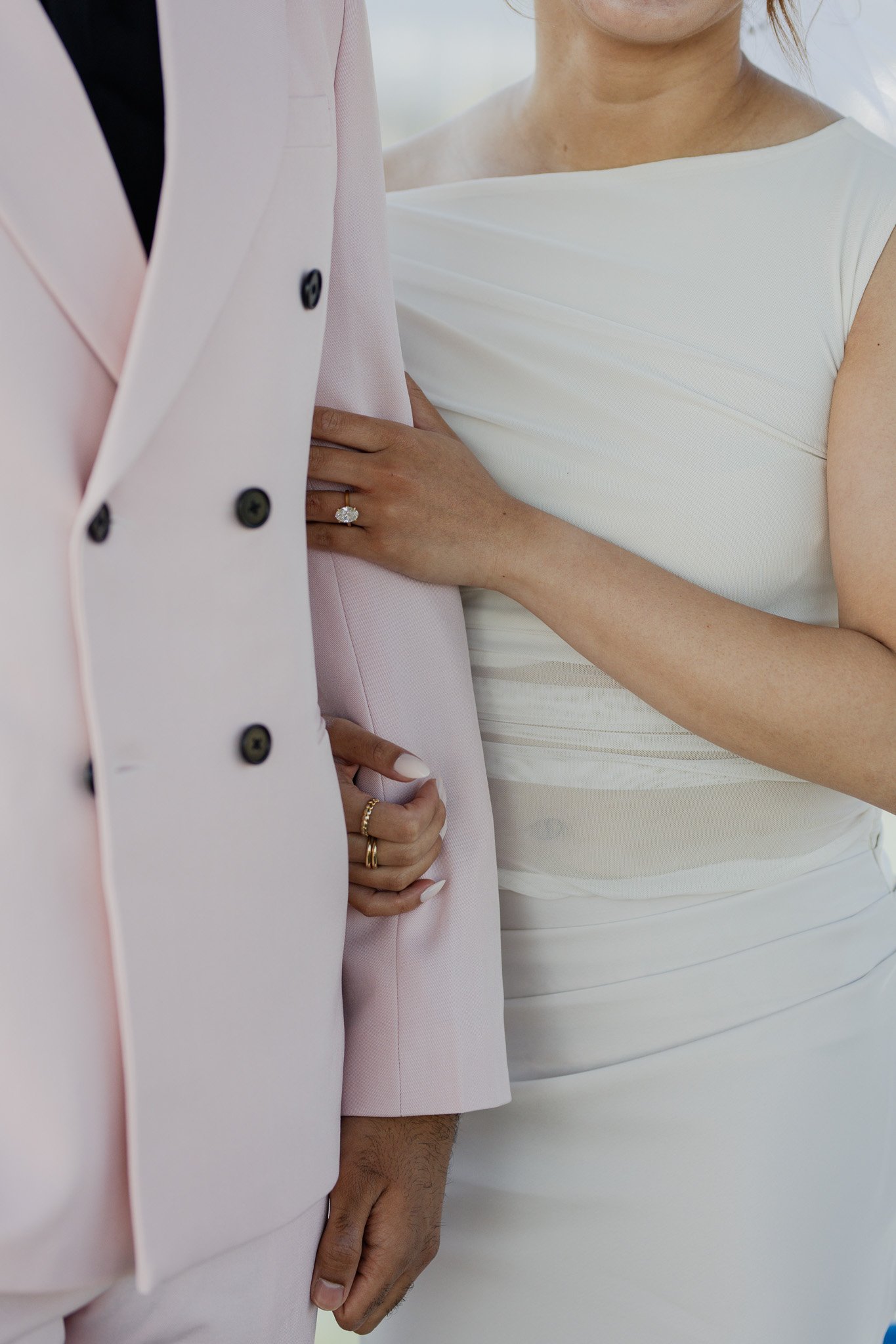 Close-up of a woman wearing a white dress with her hand resting on her waist, showing a diamond ring, standing beside a man in a pink suit with black buttons, hands visible at their sides.
