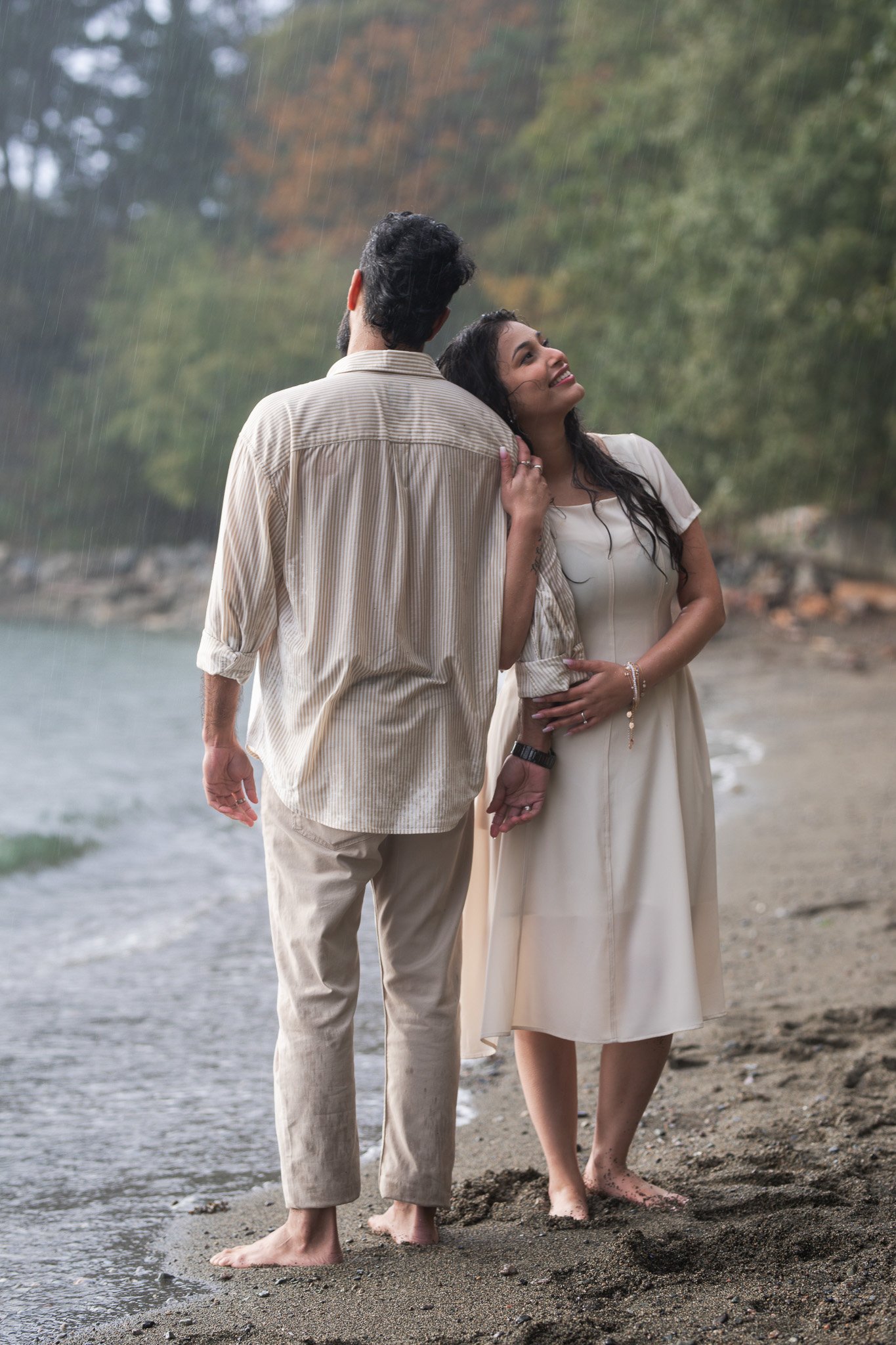 A couple holding hands on a beach during a rainstorm, embracing each other, with trees in the background.