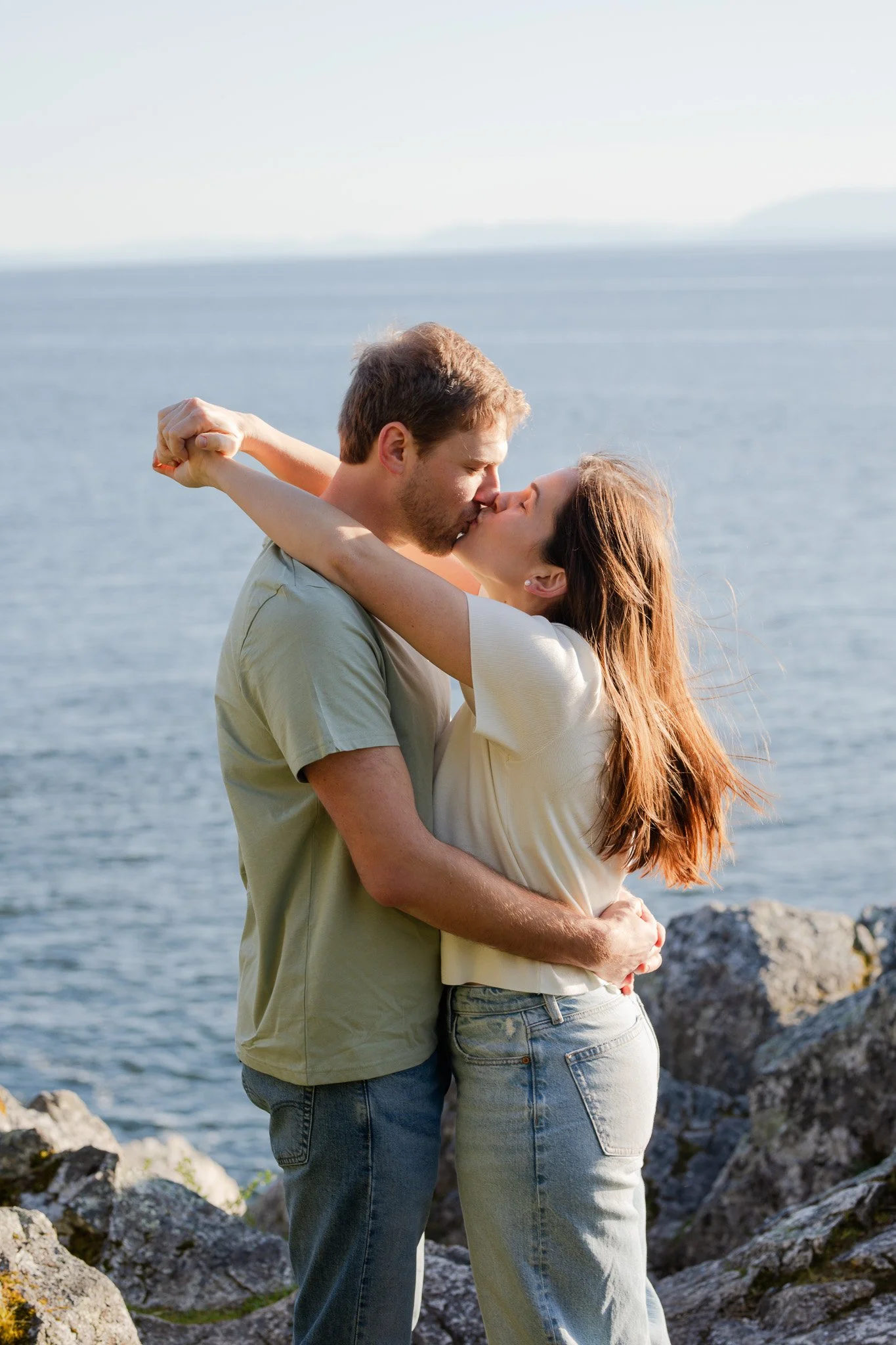 A loving couple sharing a kiss near a body of water with rocks in the foreground.