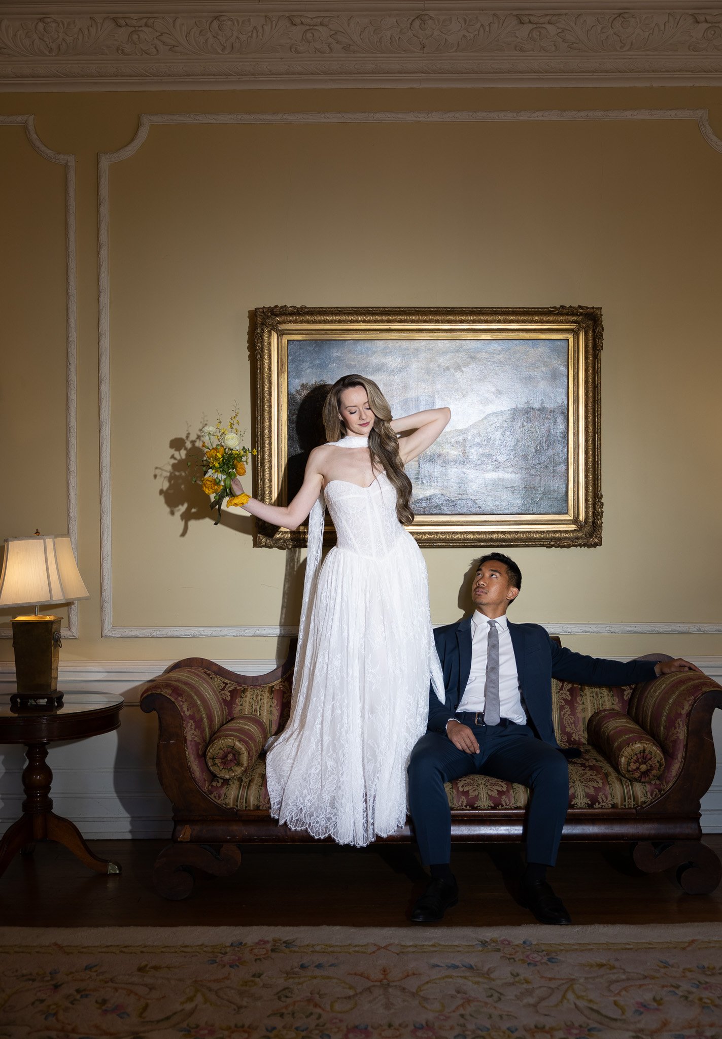 A woman in a white wedding dress holding a bouquet of flowers stands with her eyes closed, one arm behind her head, while a man in a dark suit and tie sits on a vintage sofa, looking up at her. The background features a large landscape painting, orna