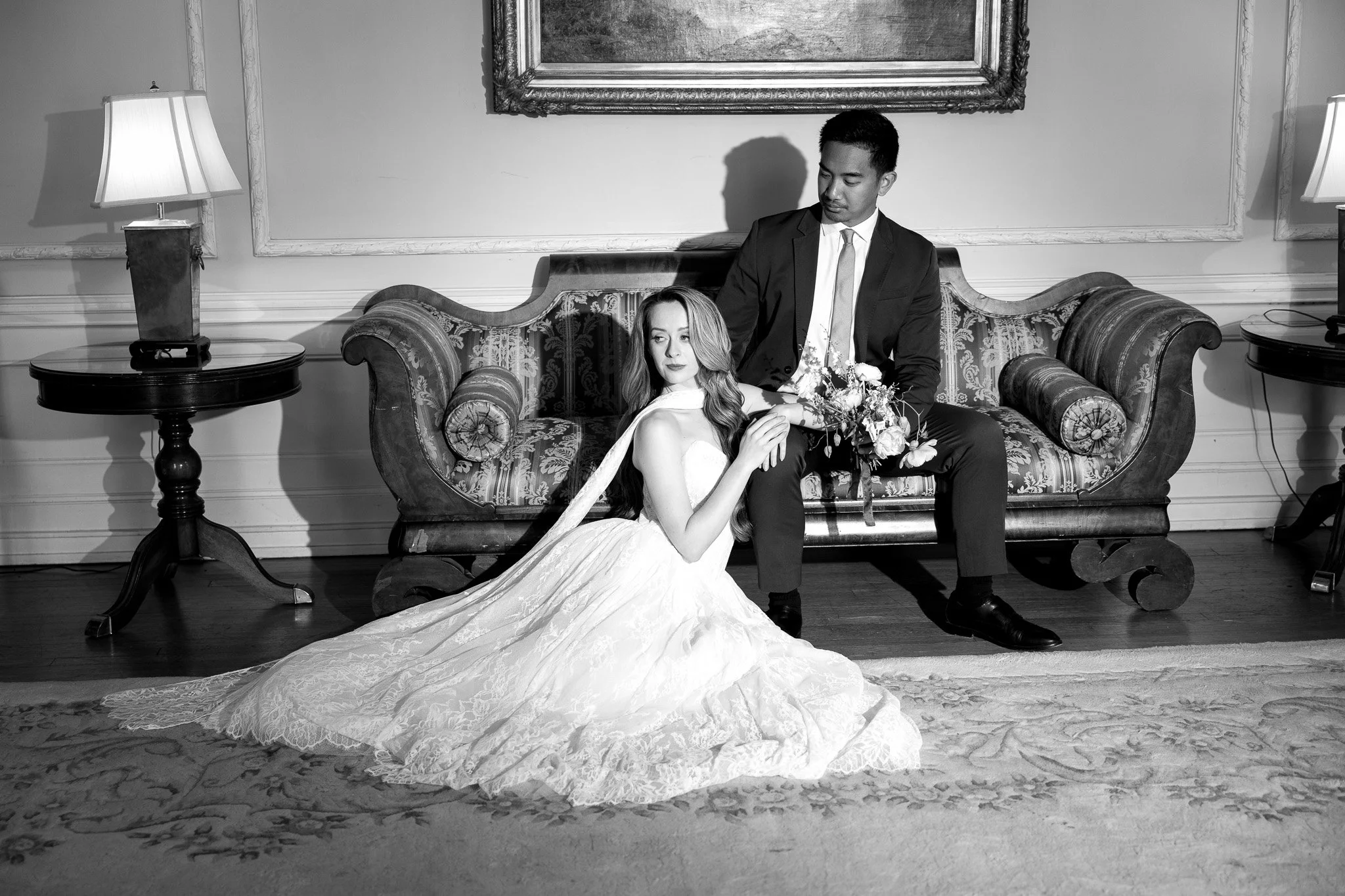 A black-and-white photo of a man in a suit sitting on a vintage sofa, holding a bouquet of flowers, with a woman in a wedding dress sitting in front of him on the floor, leaning against his legs as she looks at the camera. The setting is an elegant r