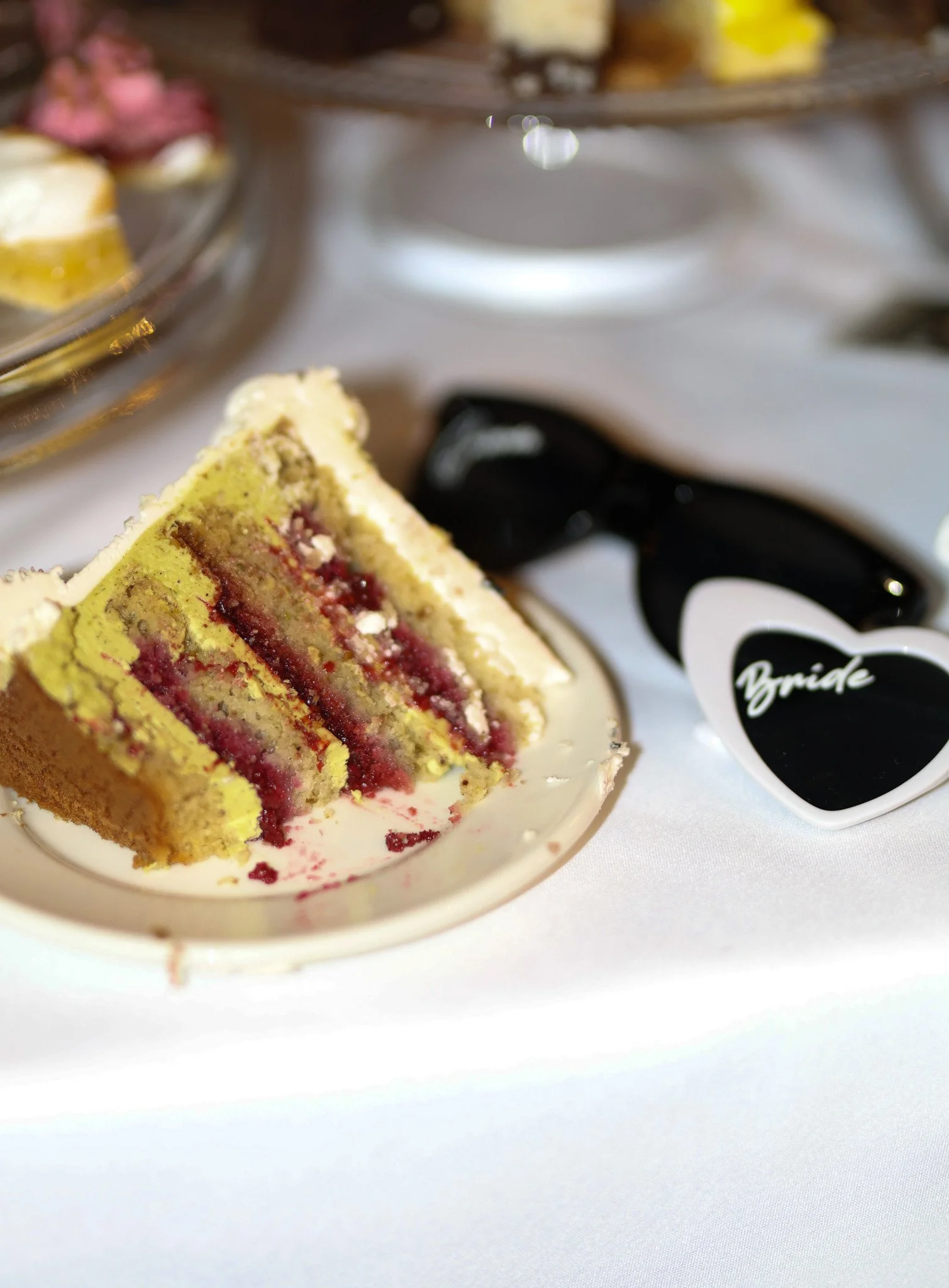 A slice of layered cake with yellow and red filling, on a white plate. In the background, a cake stand with more slices of cake and a pair of sunglasses with a sticker that says 'Bride'.