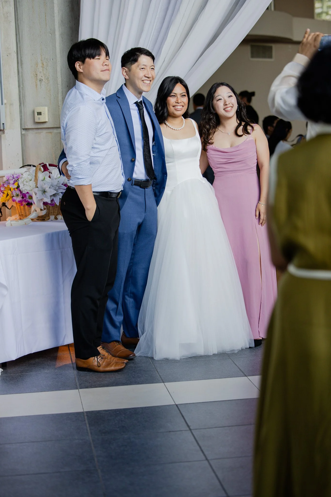Group of four people at a wedding, including the bride in a white dress and the groom in a blue suit, taking a photo together.