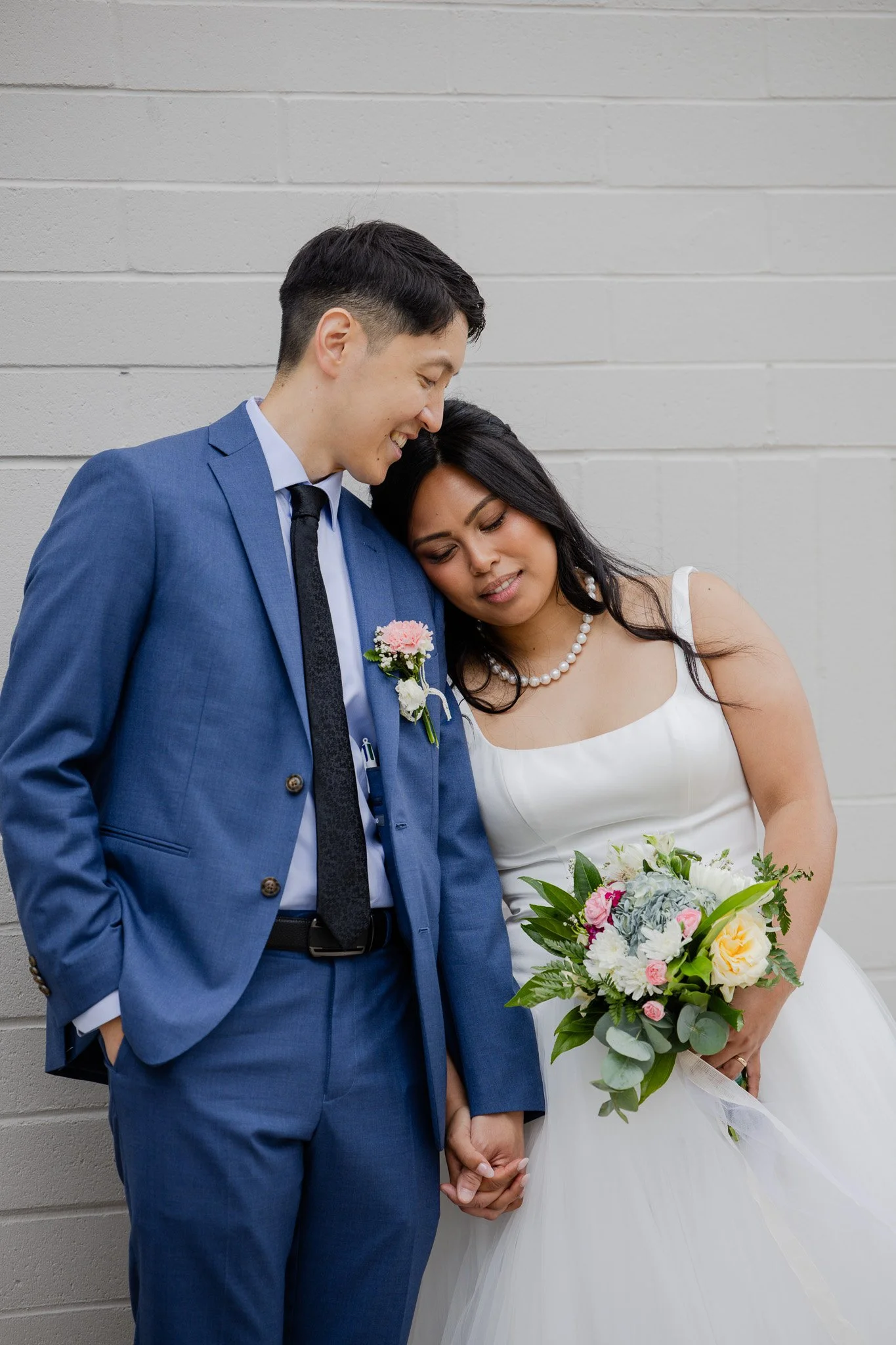 A newlywed couple standing against a white brick wall, holding hands. The groom is dressed in a blue suit with a boutonniere, and the bride in a white dress is holding a bouquet of flowers.