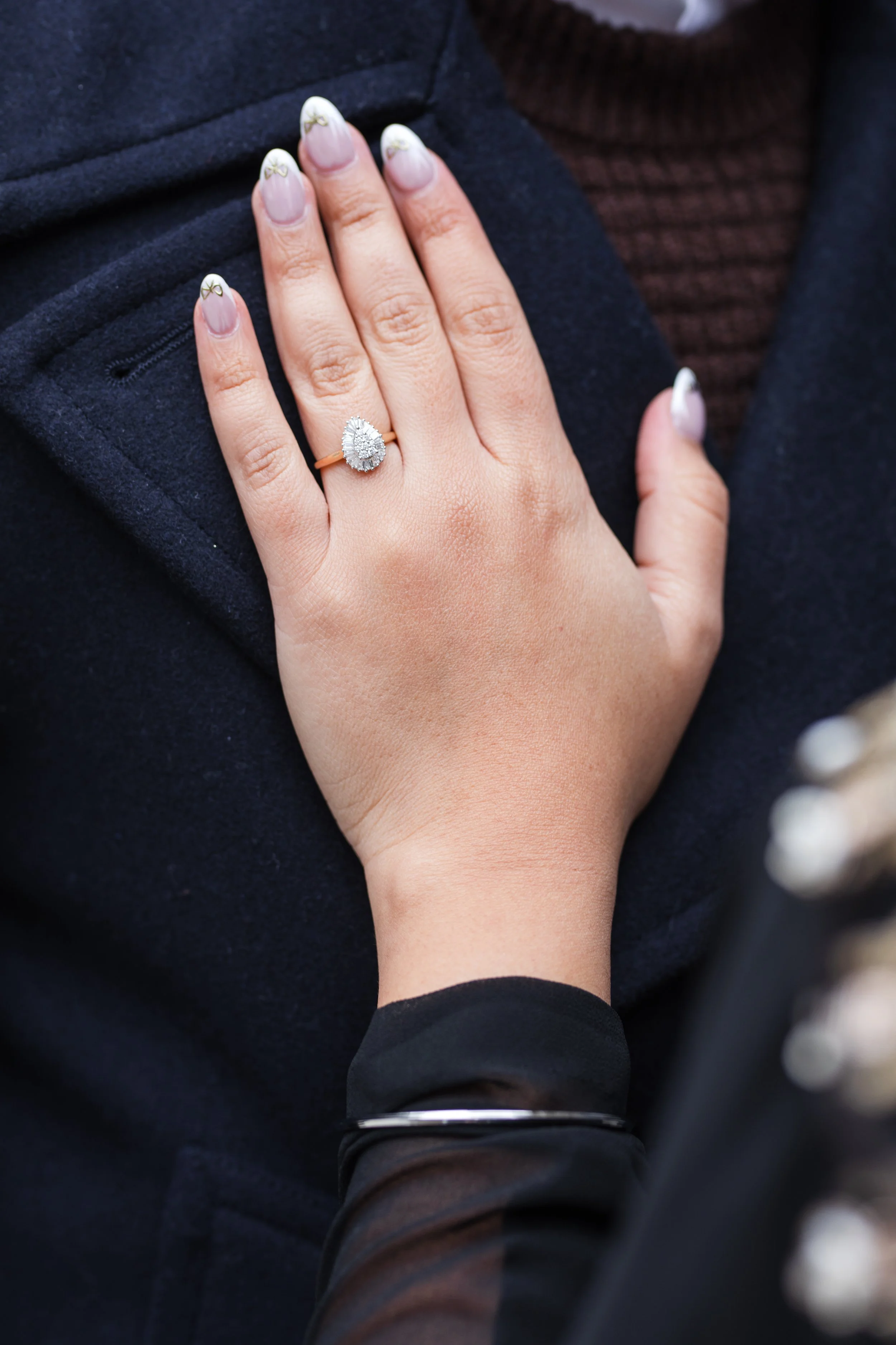 A person's hand with a ring featuring a teardrop-shaped diamond surrounded by smaller diamonds, resting on the chest with a dark jacket and a brown shirt underneath.