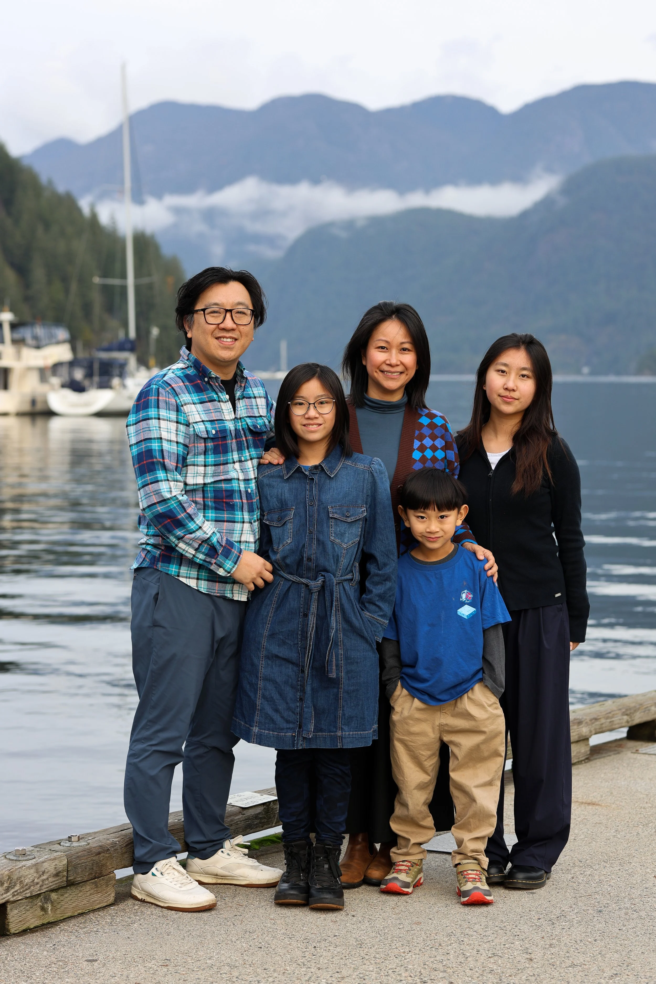 A family of six posing outdoors by a lake with mountains in the background.