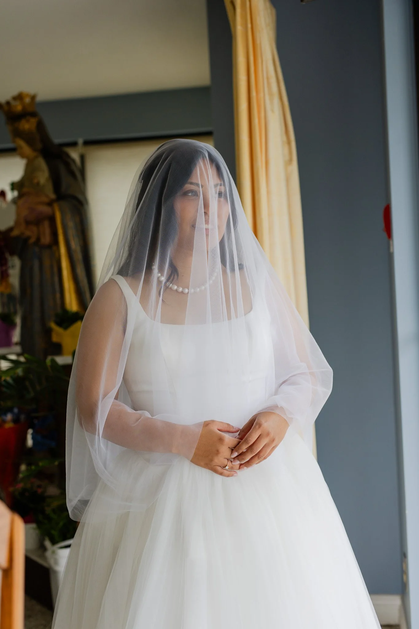 A woman in a wedding dress with a veil and pearl necklace standing indoors by a window with yellow curtains, with a statue of a religious figure and a plant in the background.
