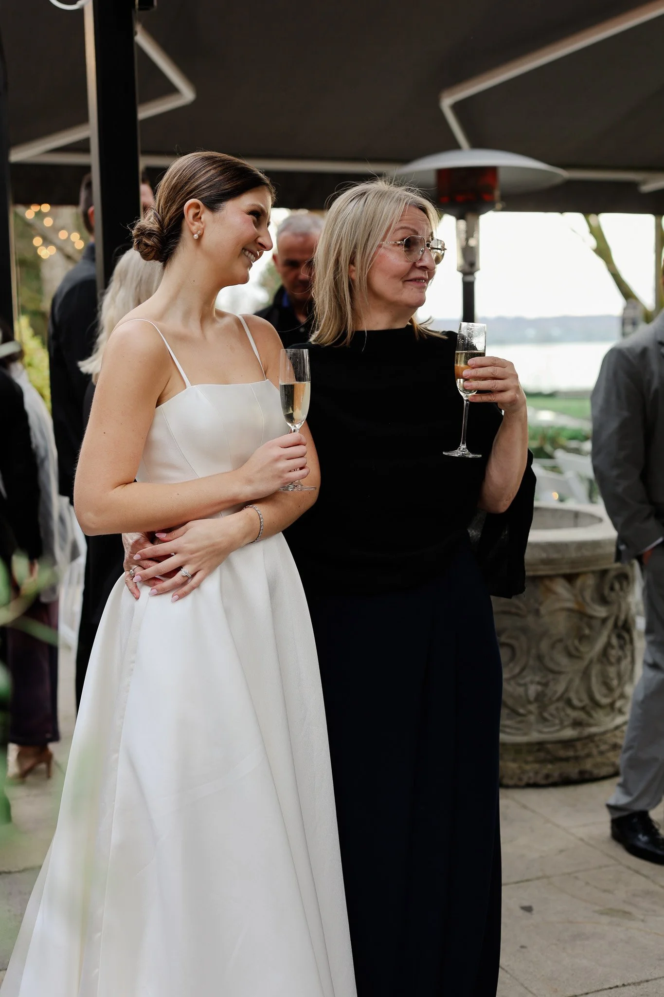 A woman in a white dress and a woman in a black dress each holding a champagne glass, at an outdoor event near a body of water with string lights and other guests in the background.