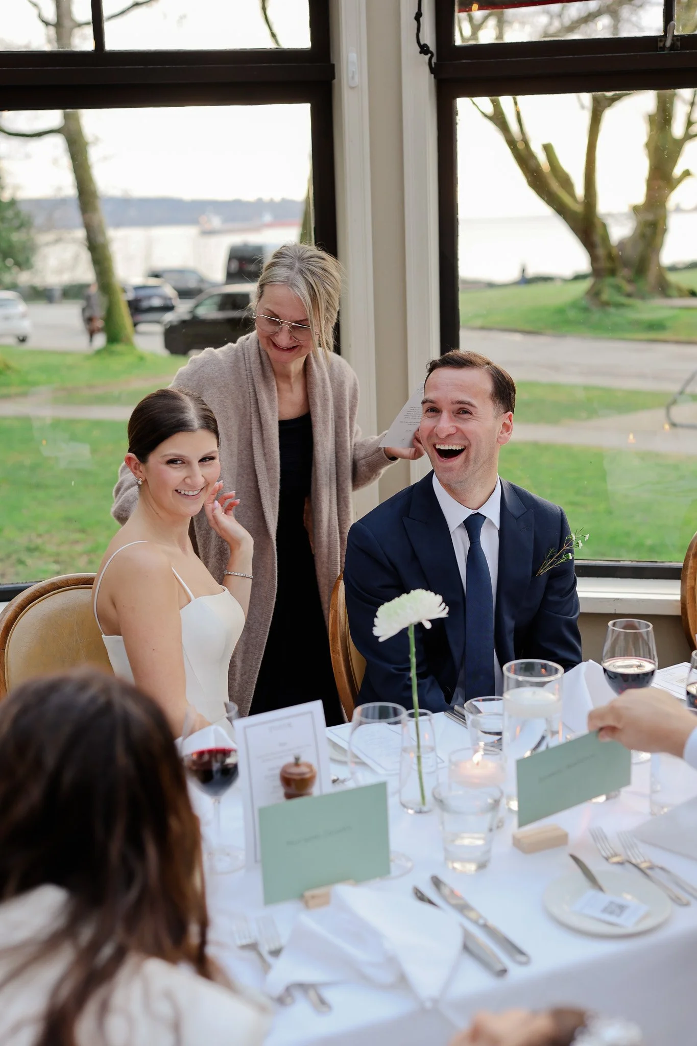 A bride and groom seated at a wedding reception table, smiling and interacting with guests. An older woman stands behind them, smiling down. The table is set with wine glasses, cutlery, menus, and a centerpiece with a white flower. Large windows show a scenic outdoor view with trees, a river, and parked cars.