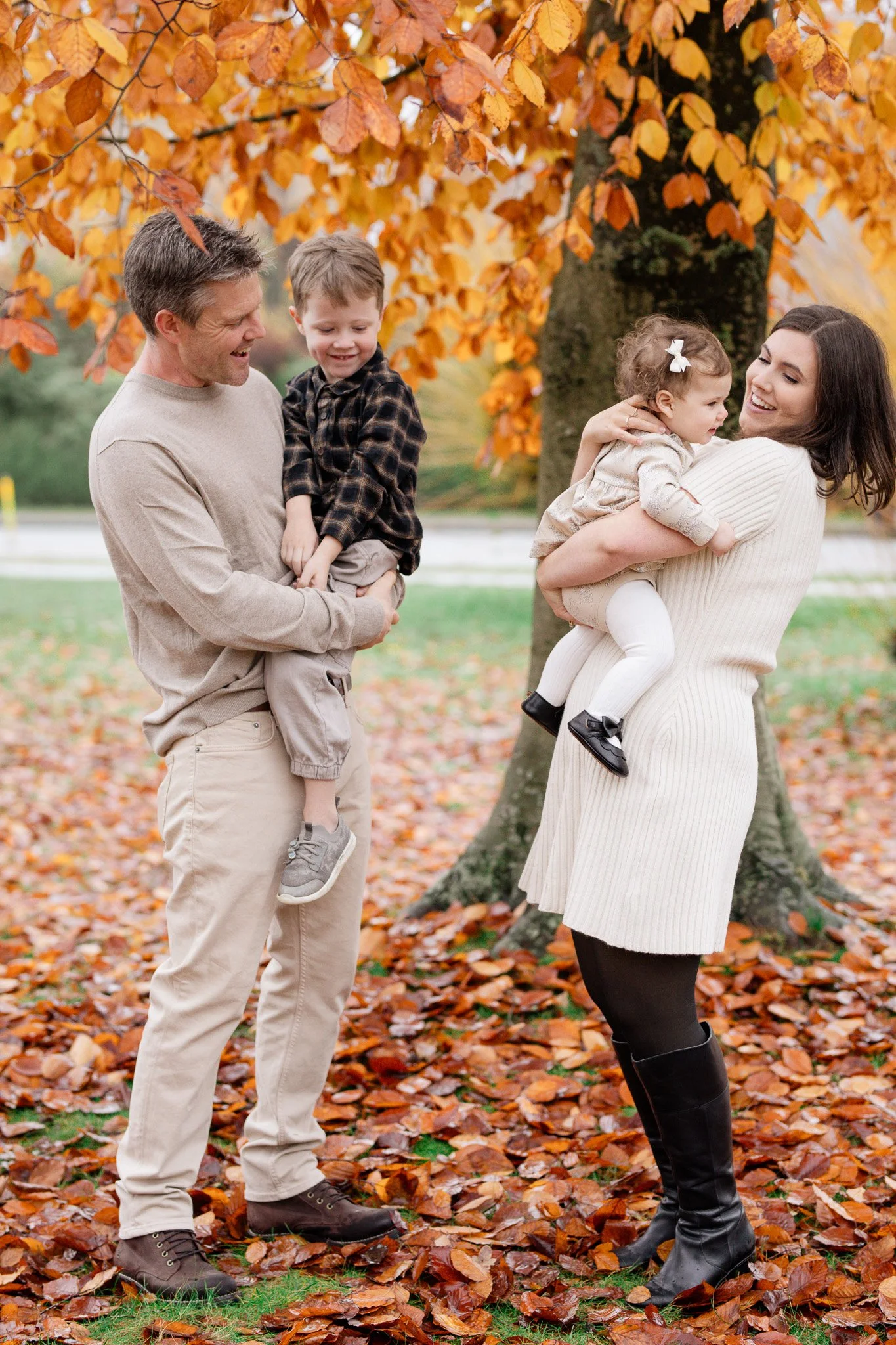 A family of four outdoors on a fall day, with orange leaves on the ground and trees in the background. The father holds a young boy, and the mother holds a young girl.