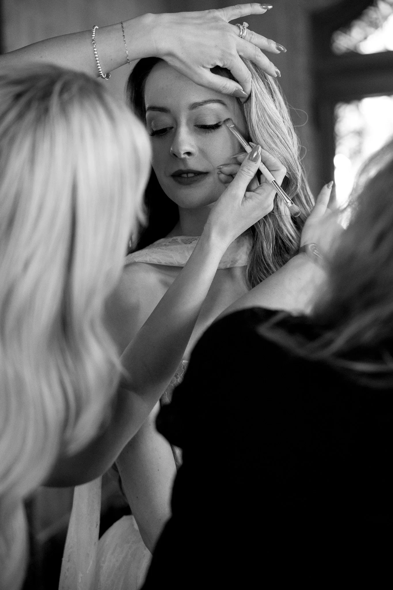 A woman with long hair getting her makeup done, with a person applying eyeliner and another person holding her head steady.