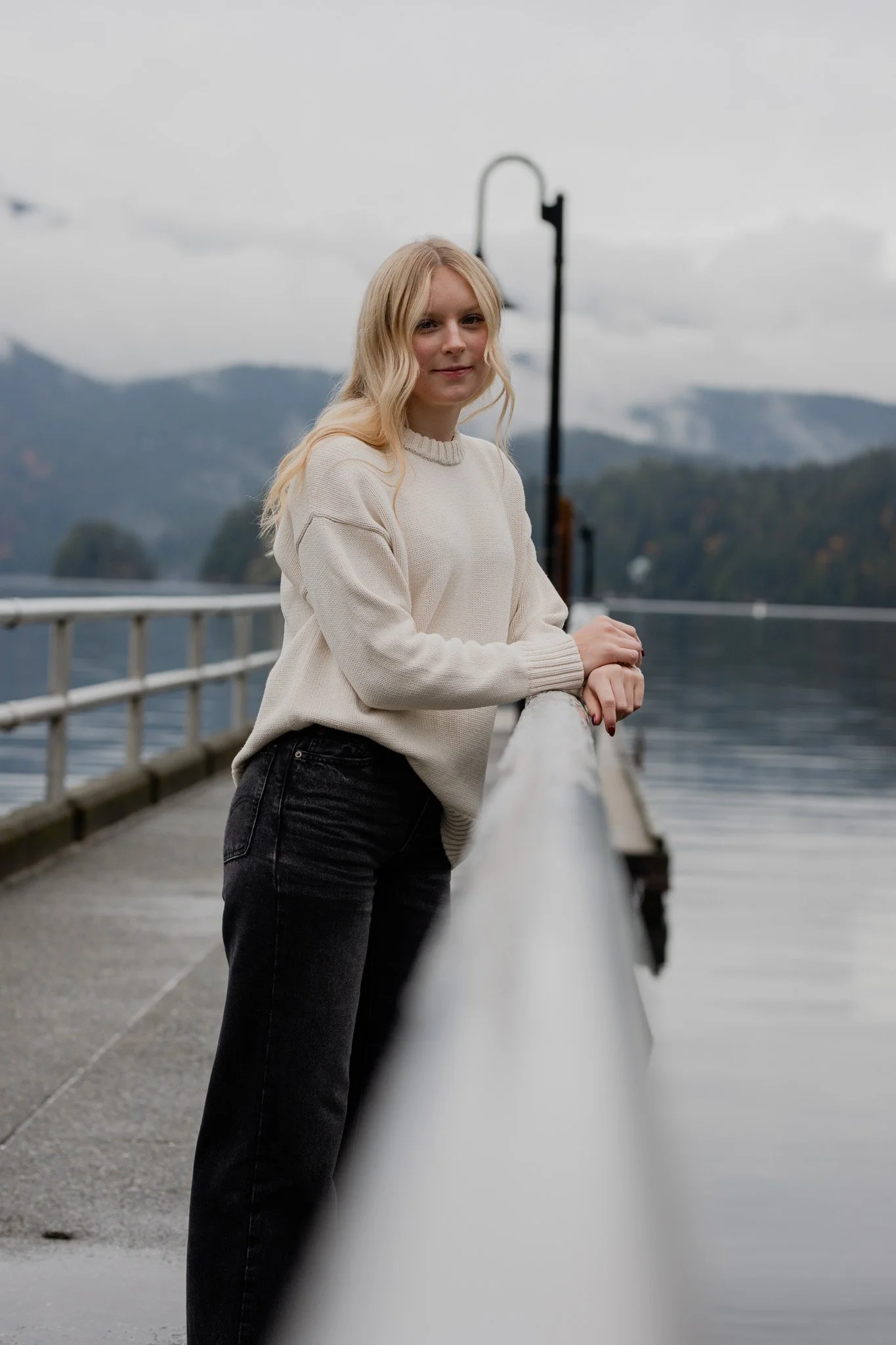 A young woman with long blonde hair stands on a dock next to a body of water, holding onto a white railing, with mountains and cloudy skies in the background.