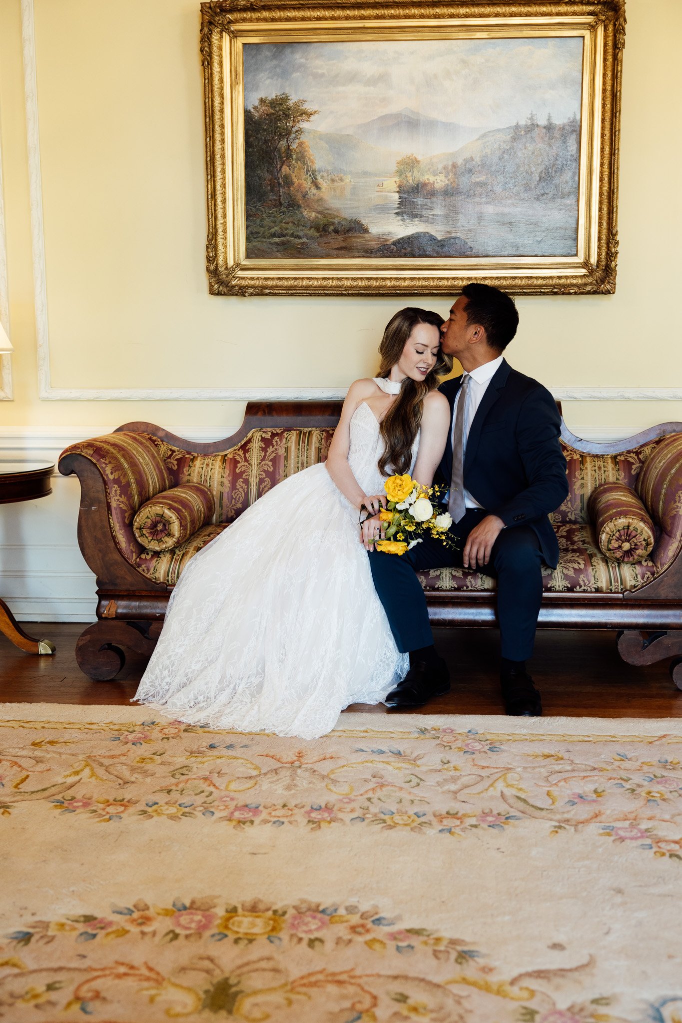 A bride and groom sitting on a vintage wooden couch in a room with yellow walls and a large landscape painting in a gold frame hanging above them. The bride is wearing a white wedding dress and holding a yellow bouquet, while the groom is wearing a d
