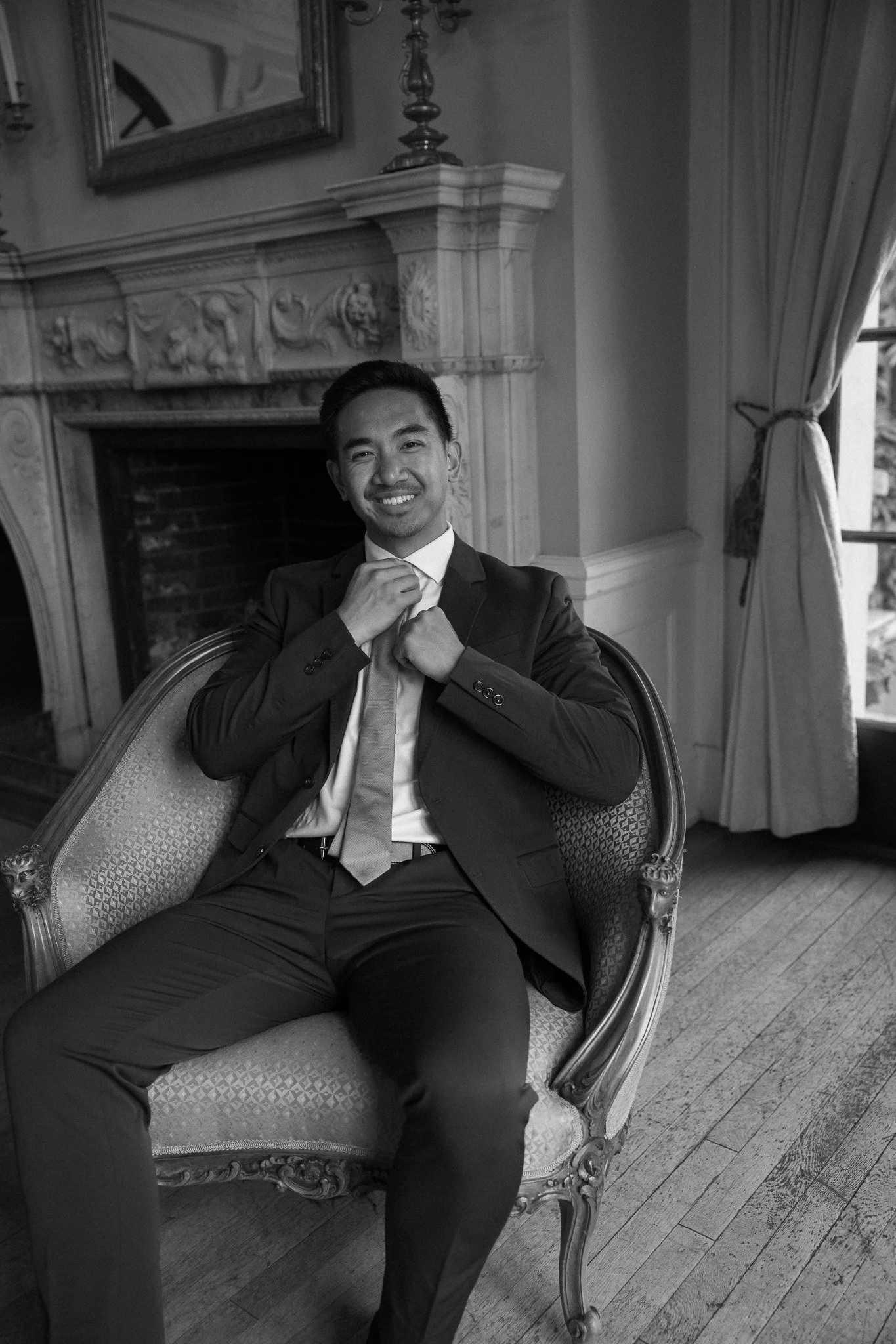 Black and white photo of a young man in a suit, sitting in an ornate vintage armchair in a room with classical decor, fireplace, mirror, curtain, and hardwood floor.