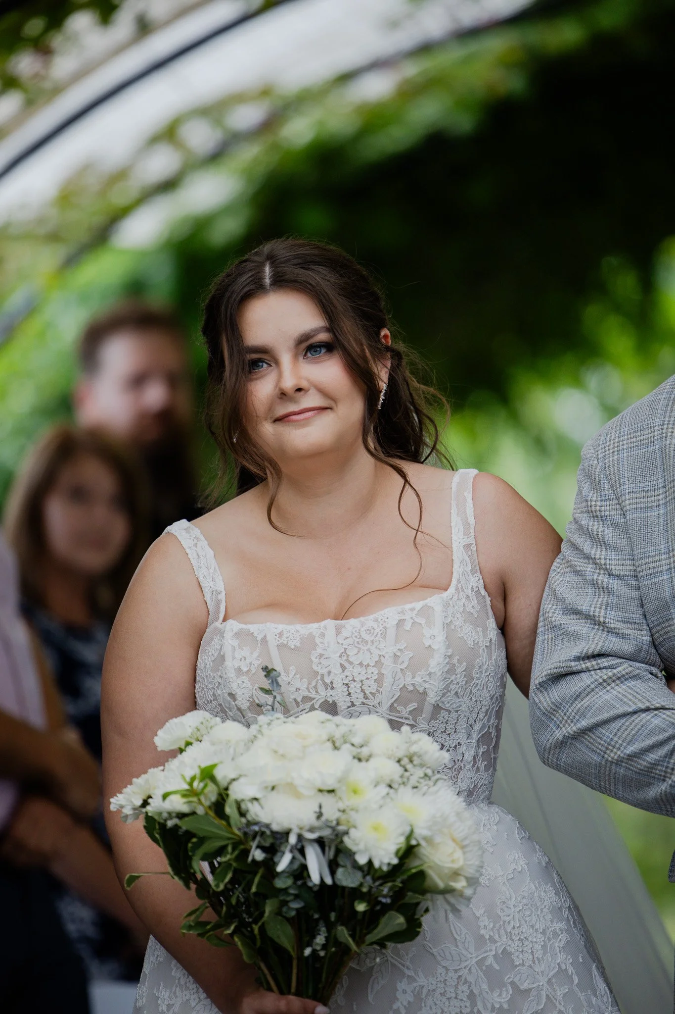 A woman with dark brown hair and blue eyes in a white lace wedding dress holding a bouquet of white flowers at a wedding ceremony.