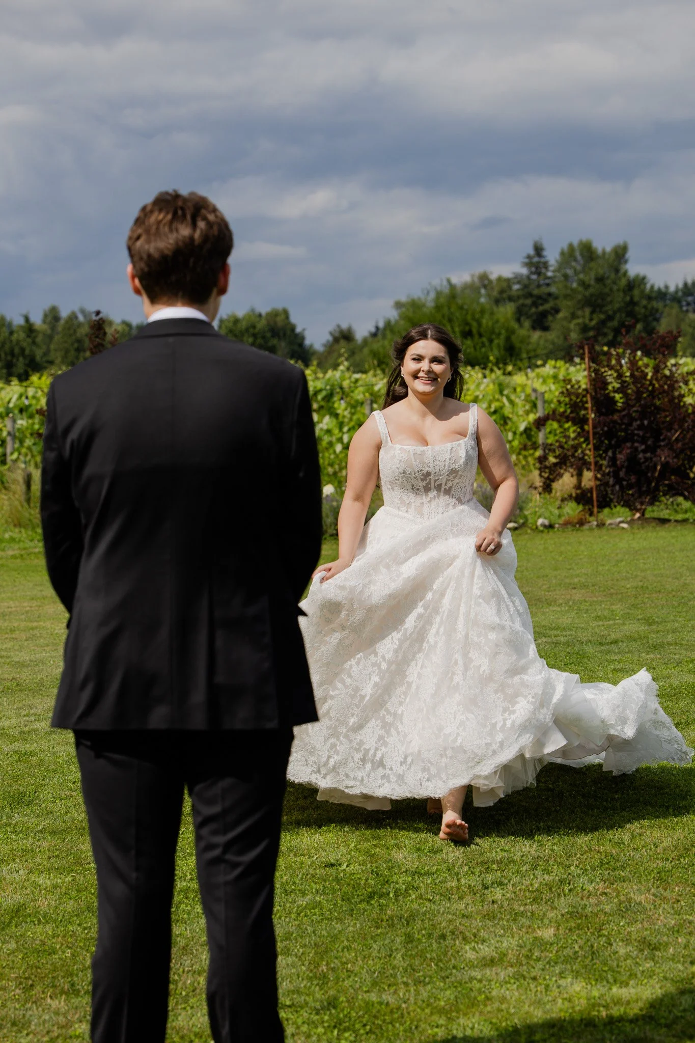 A bride in a wedding dress running outdoors towards a groom in a suit, with a background of green trees and a cloudy sky.