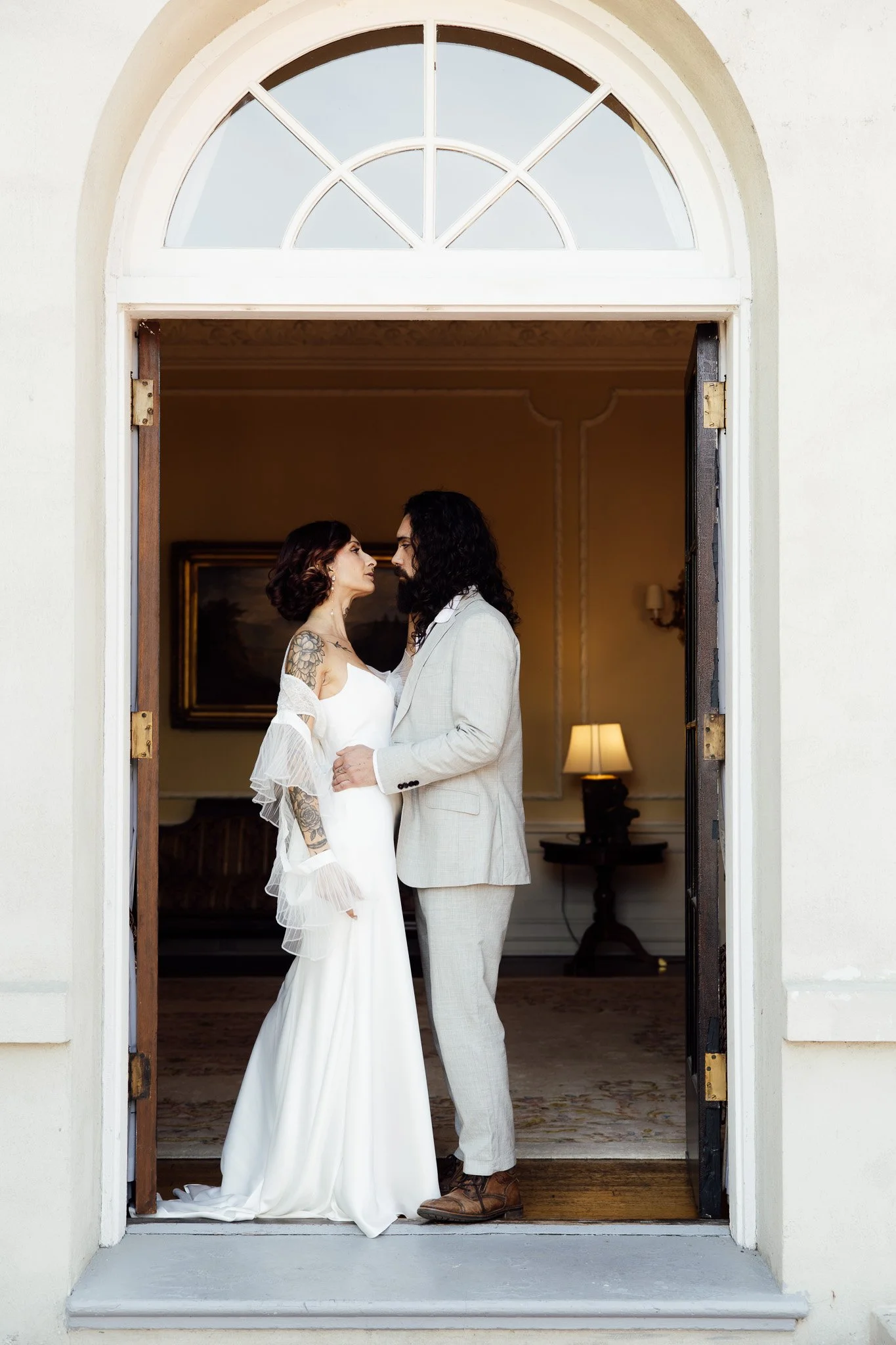 A couple in wedding attire standing face to face in a doorway of a house, about to kiss or touch foreheads, with warm indoor lighting and a painting hanging on the wall behind them.