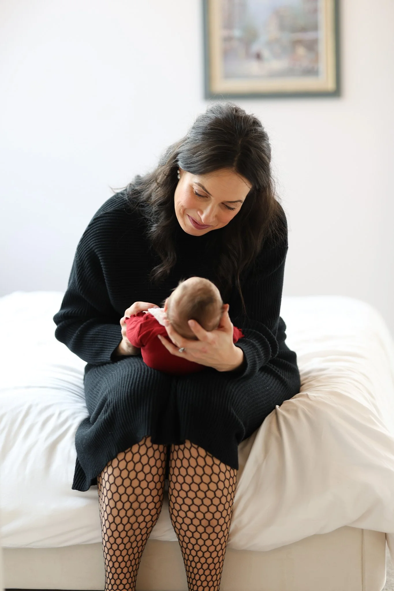 A woman in a black sweater and fishnet stockings sitting on a bed, holding a newborn baby in a red outfit and looking at the baby with a gentle expression.