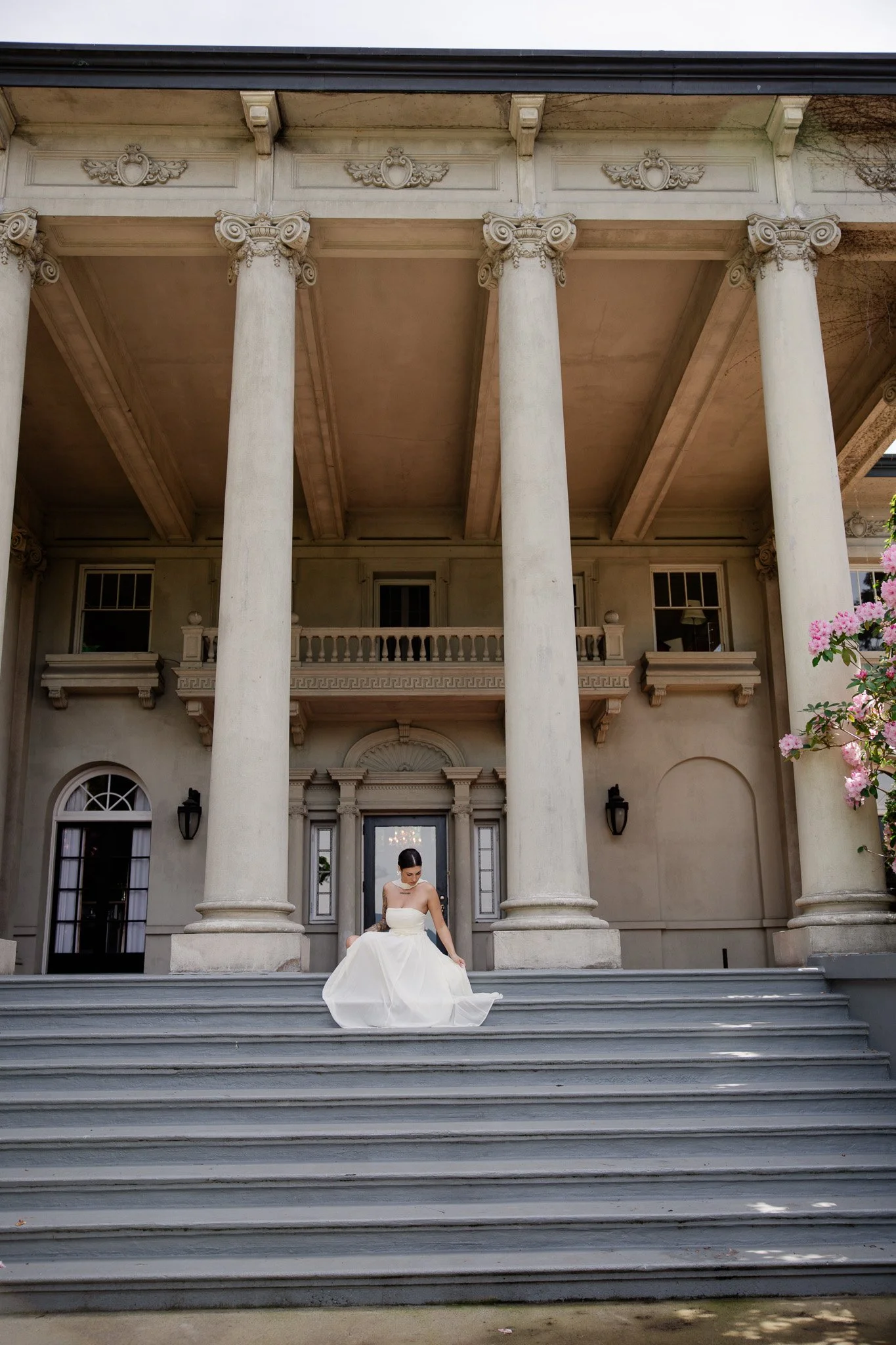 A woman in a white wedding dress sitting on the steps of a grand mansion with large columns and decorative architecture.