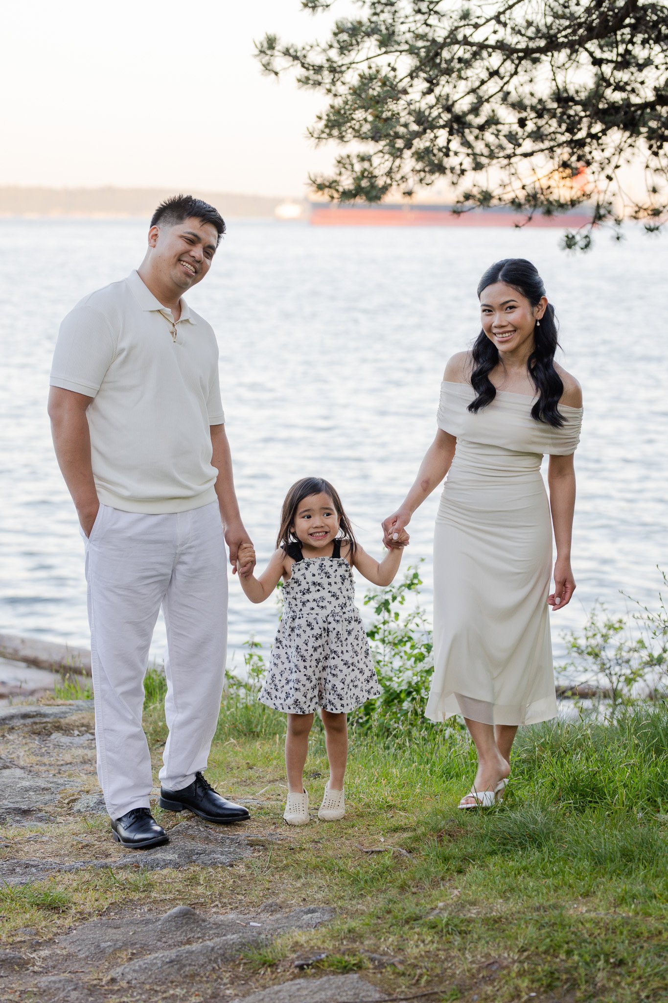 A family of four walking near a body of water, smiling and holding hands. The man is wearing a white polo shirt and white pants, the woman is in a cream off-shoulder dress, a young girl in a black and white dress, and they are outdoors with trees and