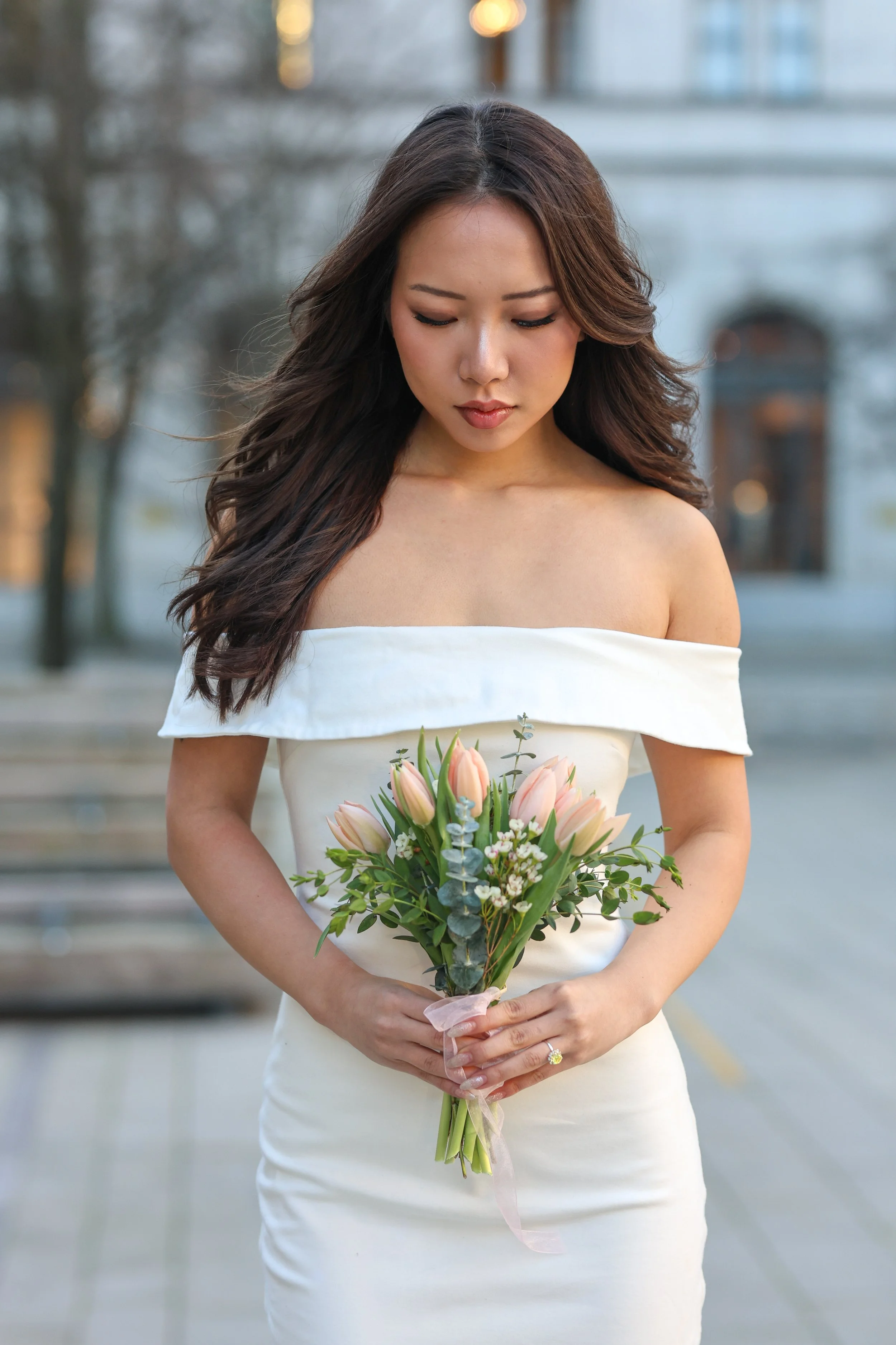 A woman in a white off-shoulder dress holding a bouquet of pink tulips and greenery, standing outdoors.