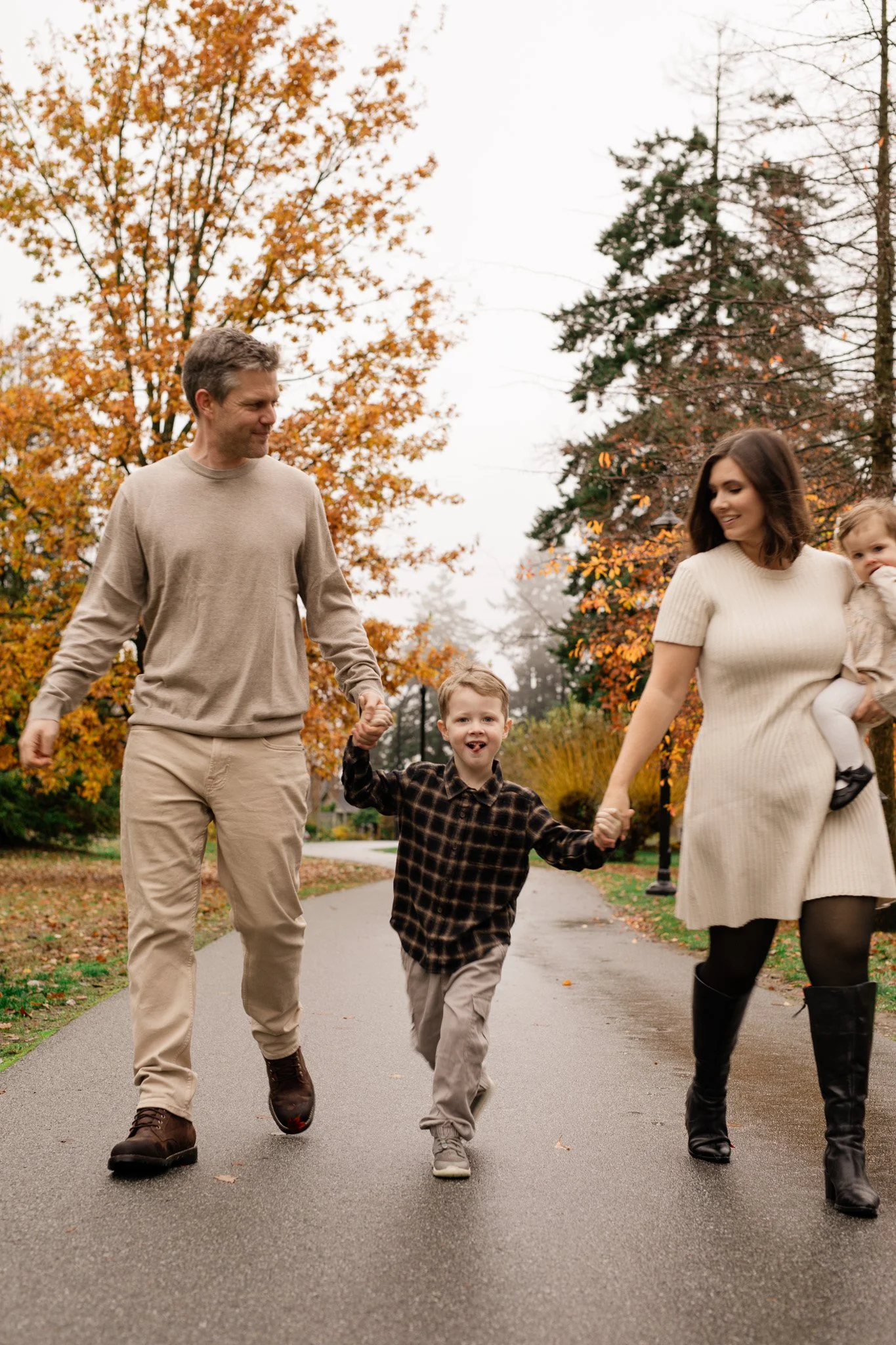 A family of four walking on a park path during fall. The father and mother are holding hands with their children, a son in the middle and a girl in the mother's arms. The trees have vibrant orange and green leaves.