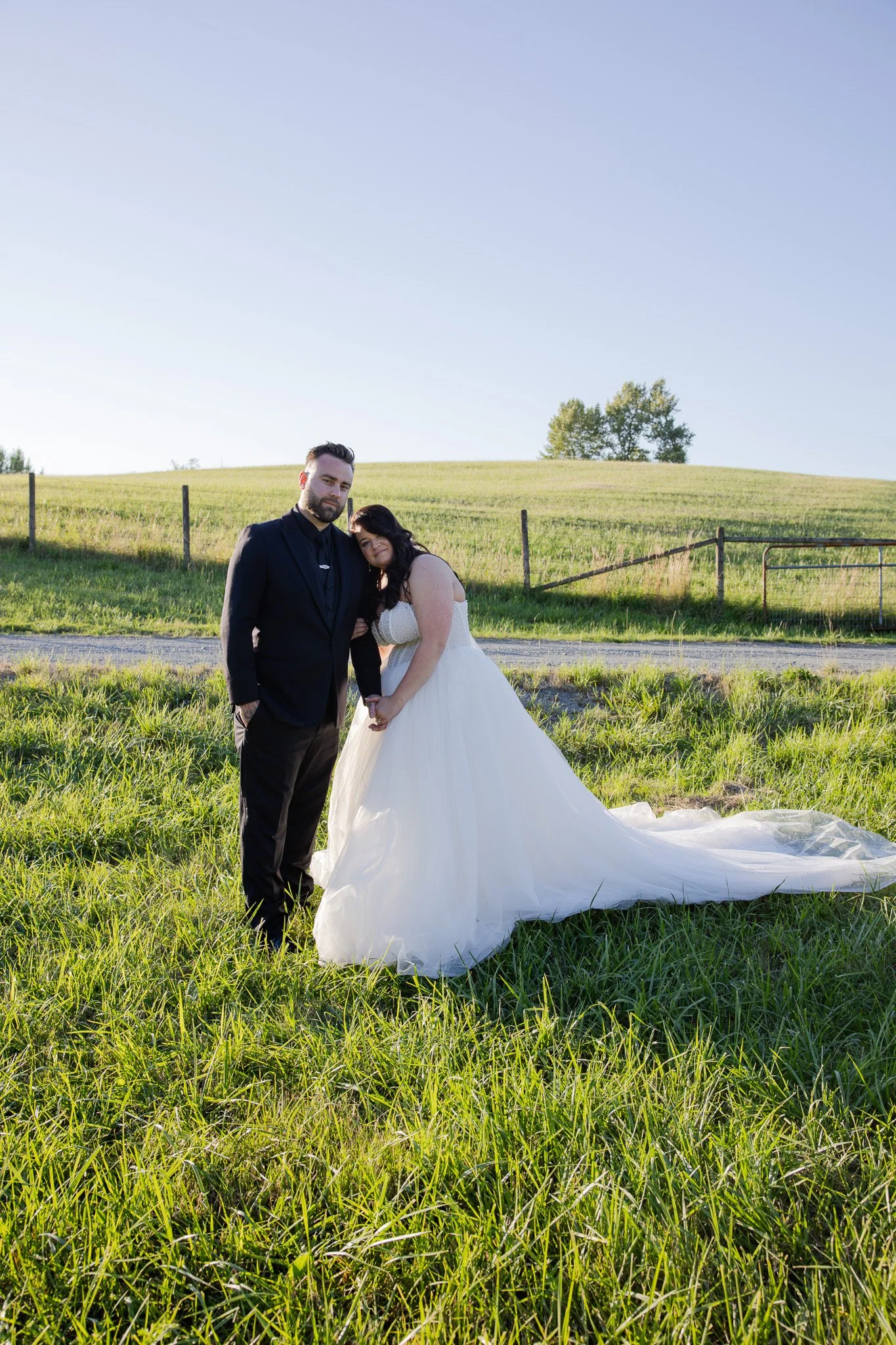 A man and woman in wedding attire holding hands outdoors in a grassy field with a fence and trees in the background.