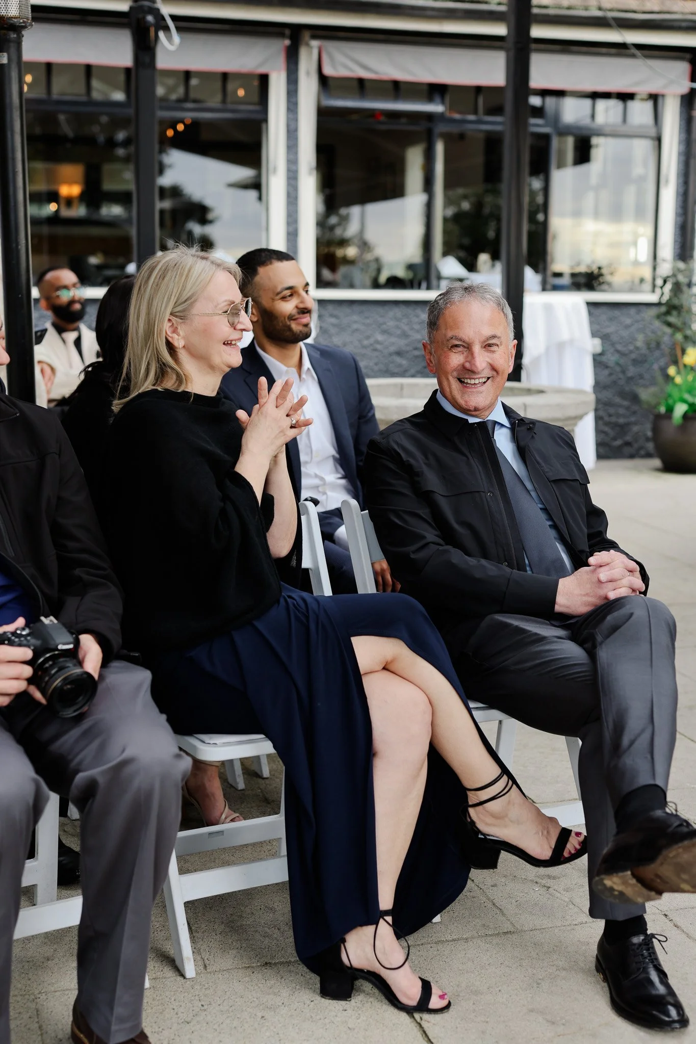 People seated outside and smiling at an event, with a woman in a black dress and high heels, an older man in a suit, and a man in a suit behind them.