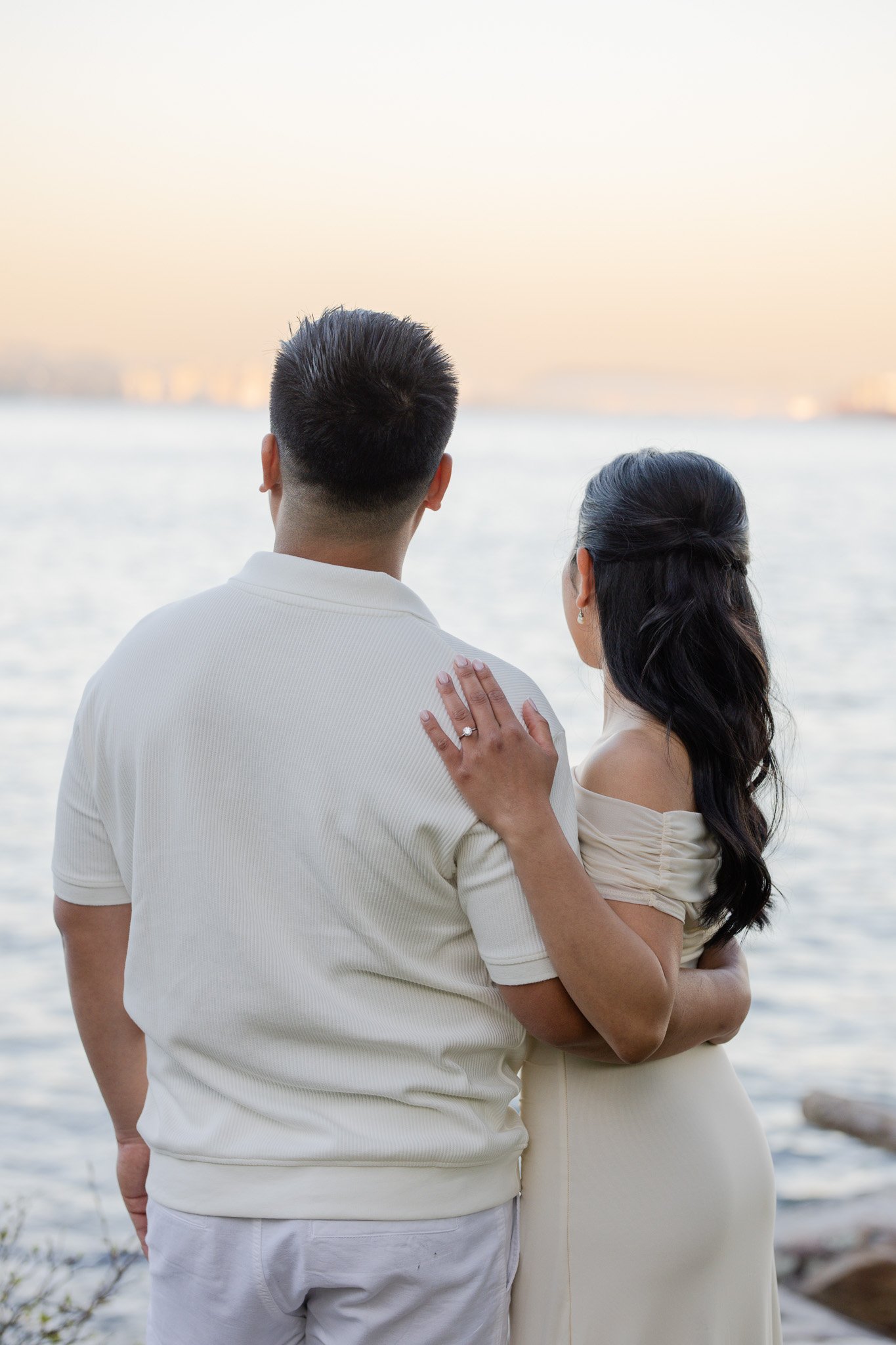 A couple stands closely together by the water, with the woman resting her hand on the man's shoulder, overlooking a calm body of water with a distant shoreline at sunset.