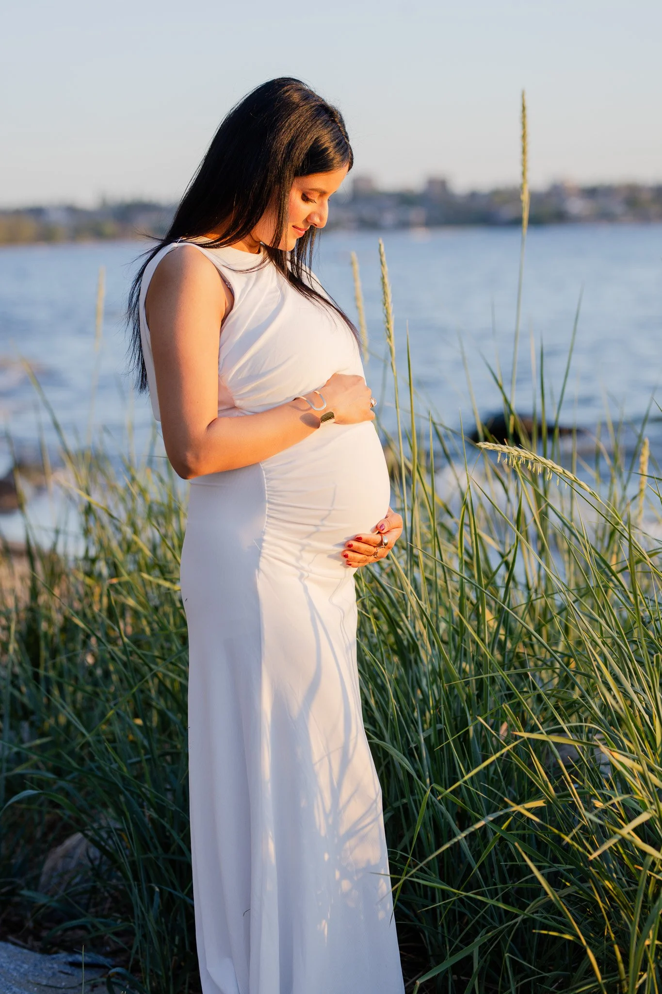 A pregnant woman standing outdoors near water, wearing a white sleeveless dress, with her hands on her belly, looking down with a peaceful expression.