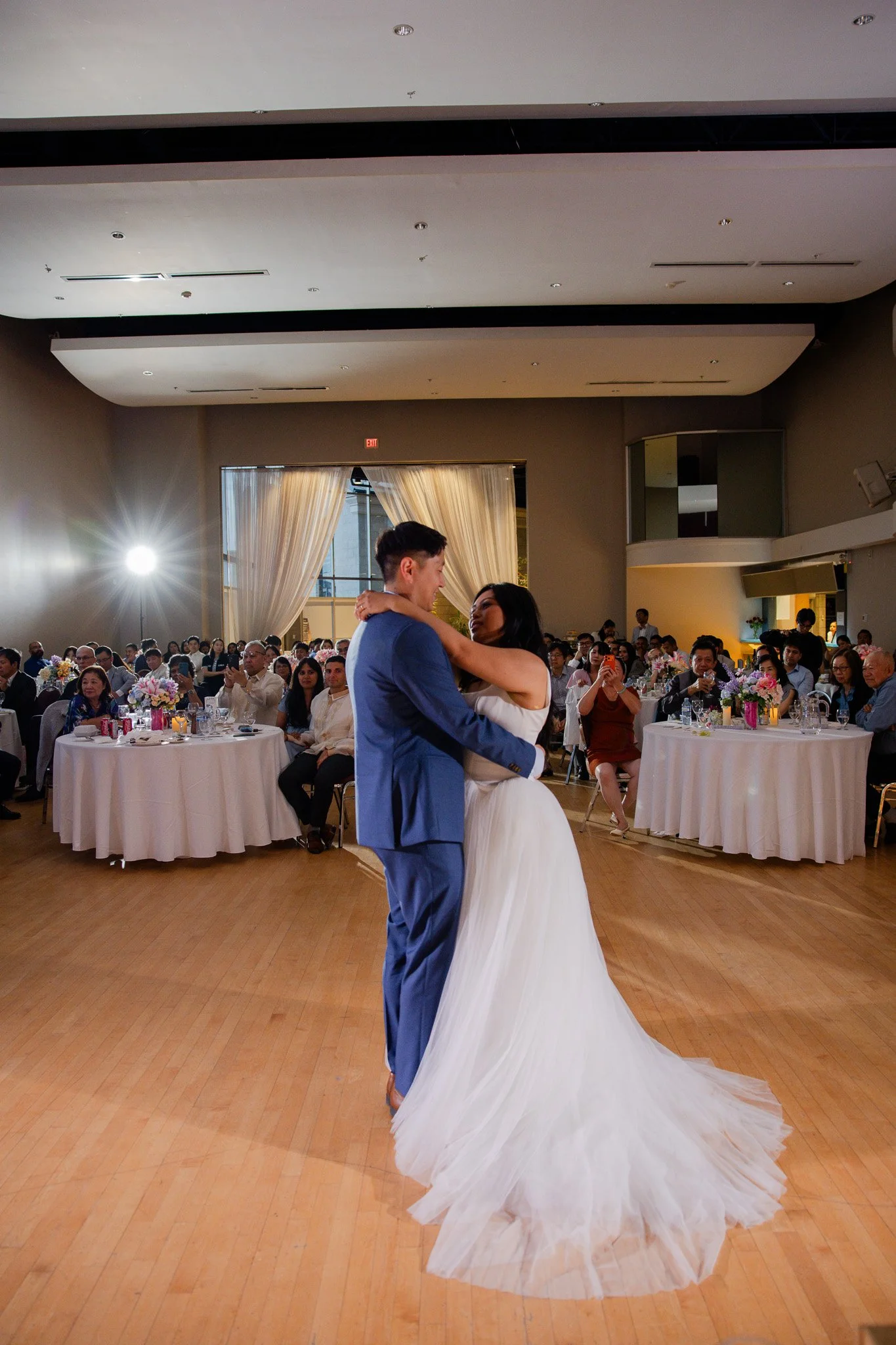 A bride and groom are dancing at their wedding reception in a banquet hall. The bride is wearing a white wedding gown, and the groom is in a blue suit. Guests seated at round tables with floral centerpieces watch and take photos.