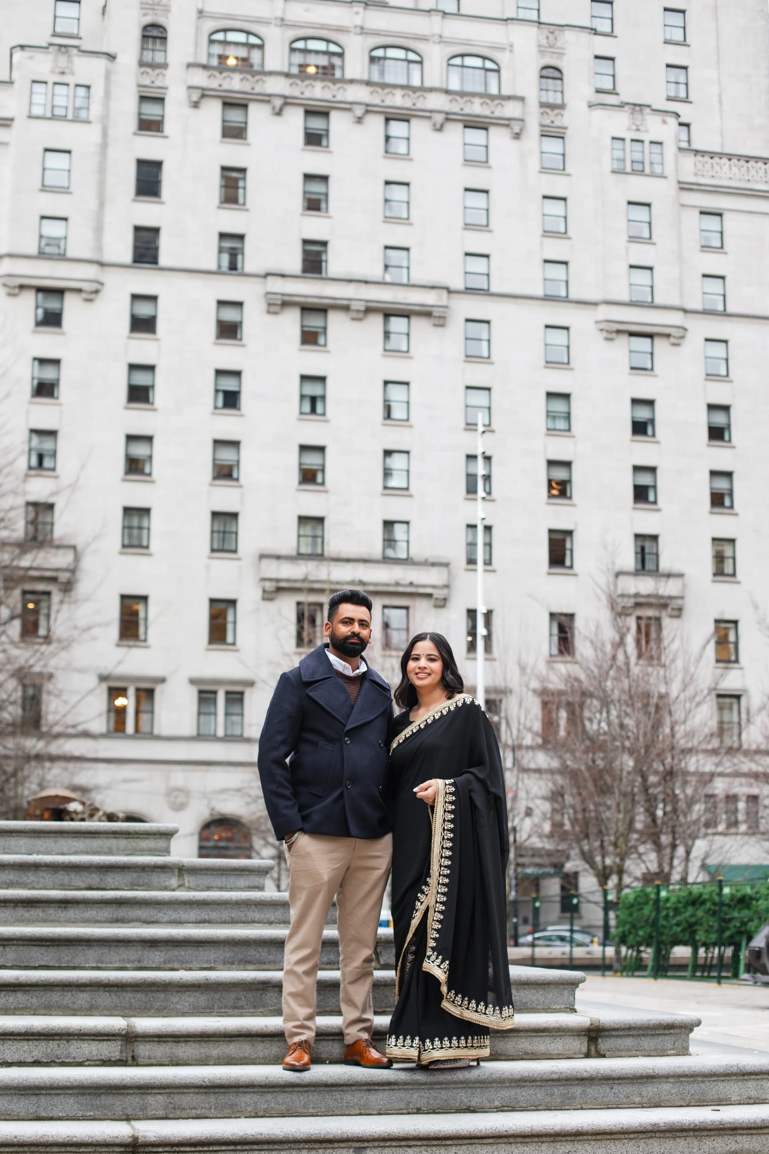 A man and woman standing together on steps in front of a tall, beige building with many windows. The woman wears a black sari with gold embroidery, and the man is dressed in a navy coat and beige pants.