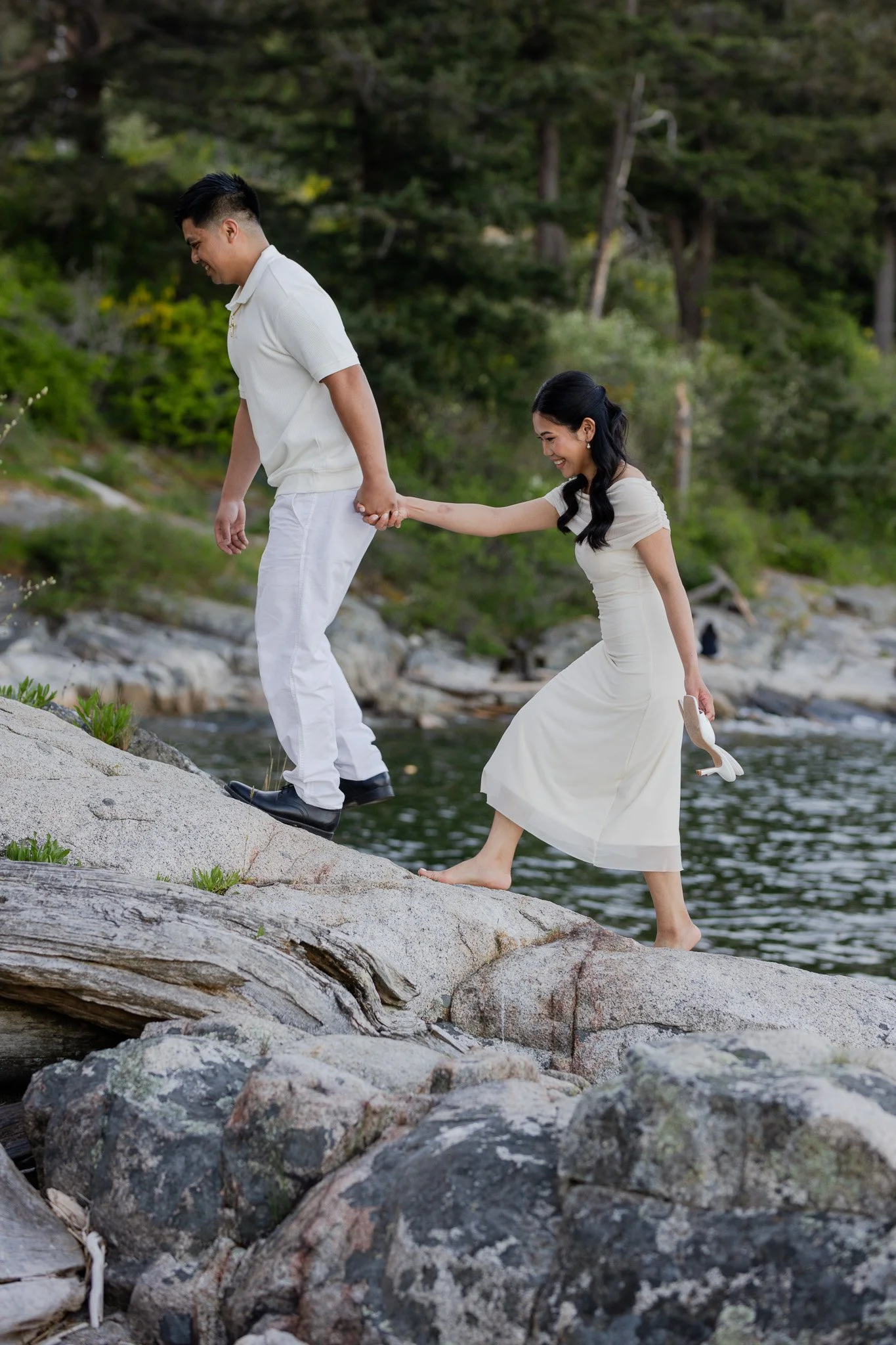 A couple walking carefully on rocks by a river, with the woman holding the man's hand for balance and both smiling.