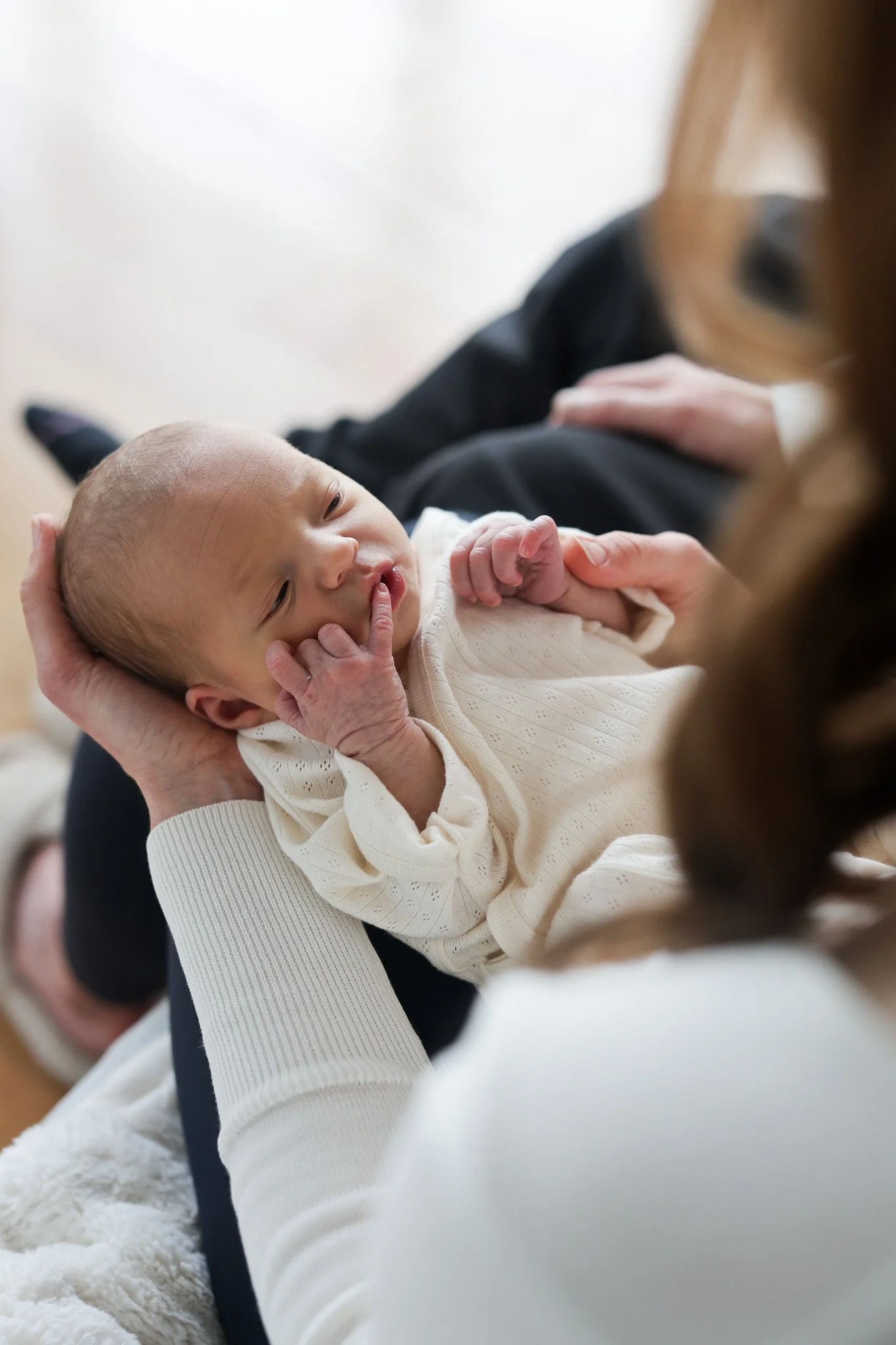 Close-up of a baby with a bald head lying in someone's arms, holding a finger near their mouth, with adult person partially visible and wearing a dark top in the background.