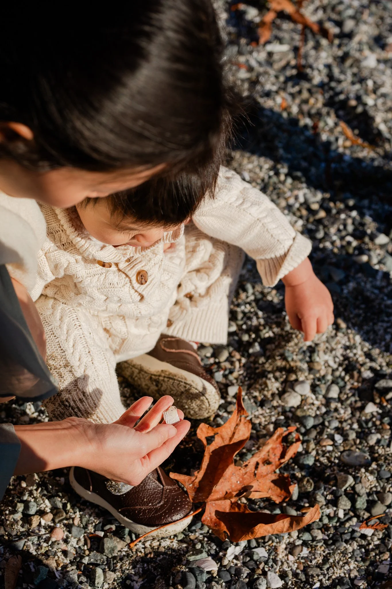 Adult and two children examining fallen autumn leaves on a gravelly ground.