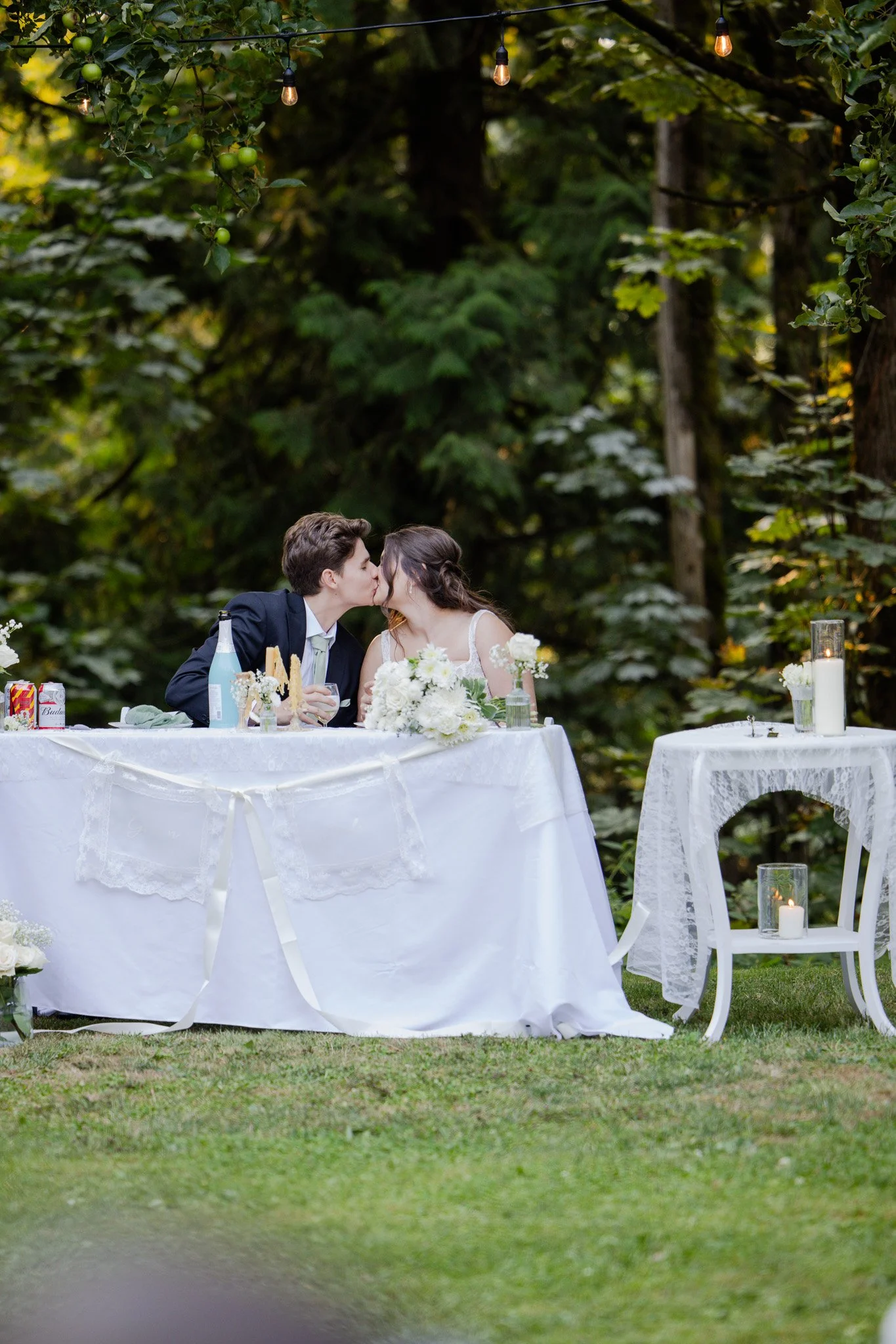 A bride and groom sharing a kiss at their outdoor wedding reception, seated at a table decorated with white flowers and candles, surrounded by green trees and string lights.