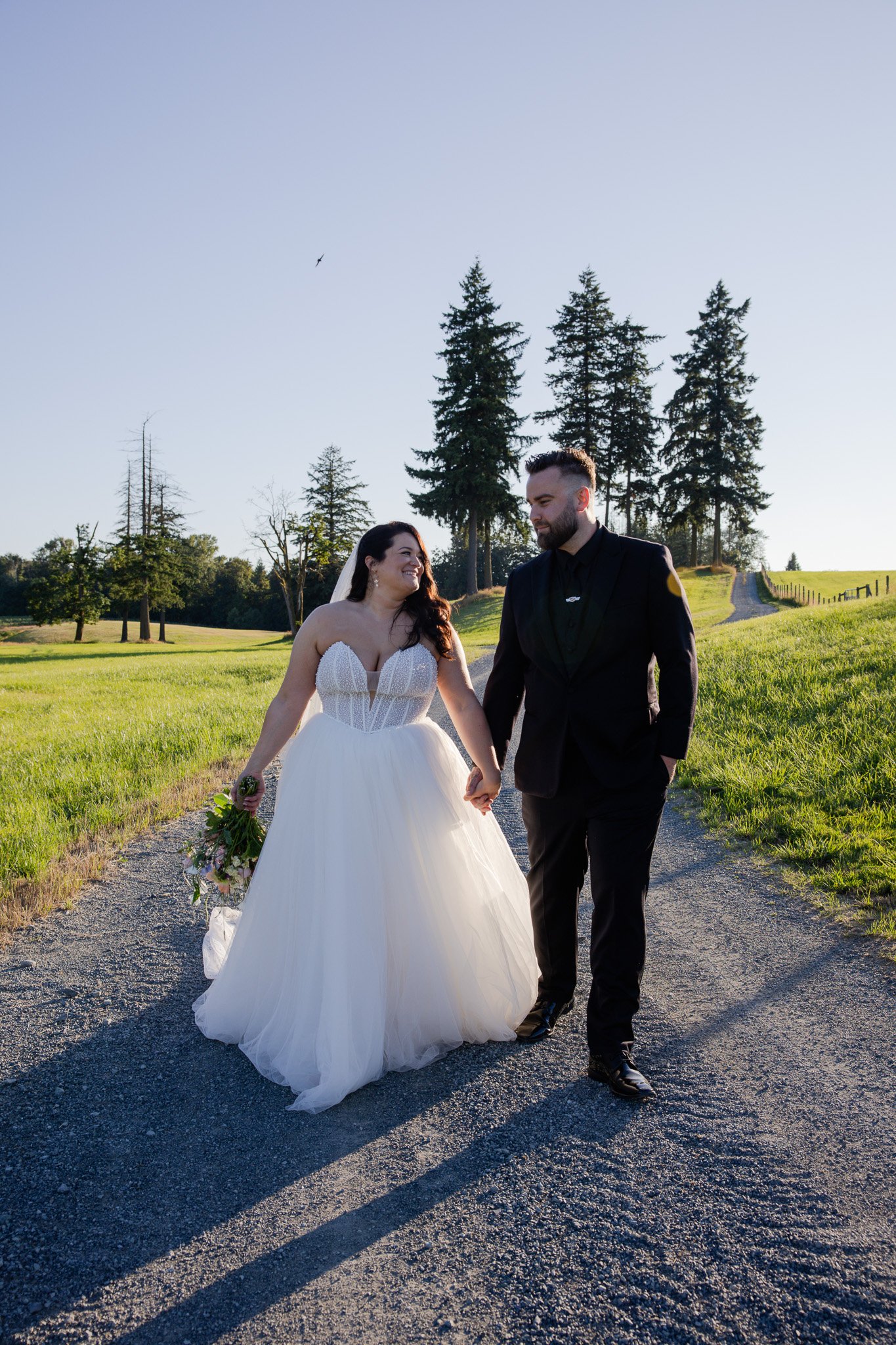 A bride and groom walking hand in hand on a dirt path in a green field with tall trees in the background, under a clear blue sky, during sunset.