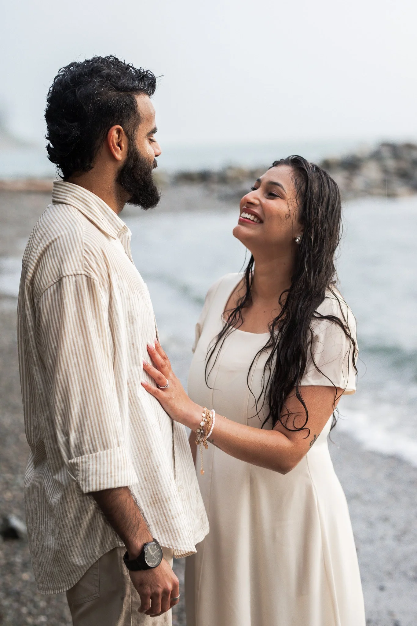 A couple standing on the beach, smiling and looking at each other, with the ocean in the background.