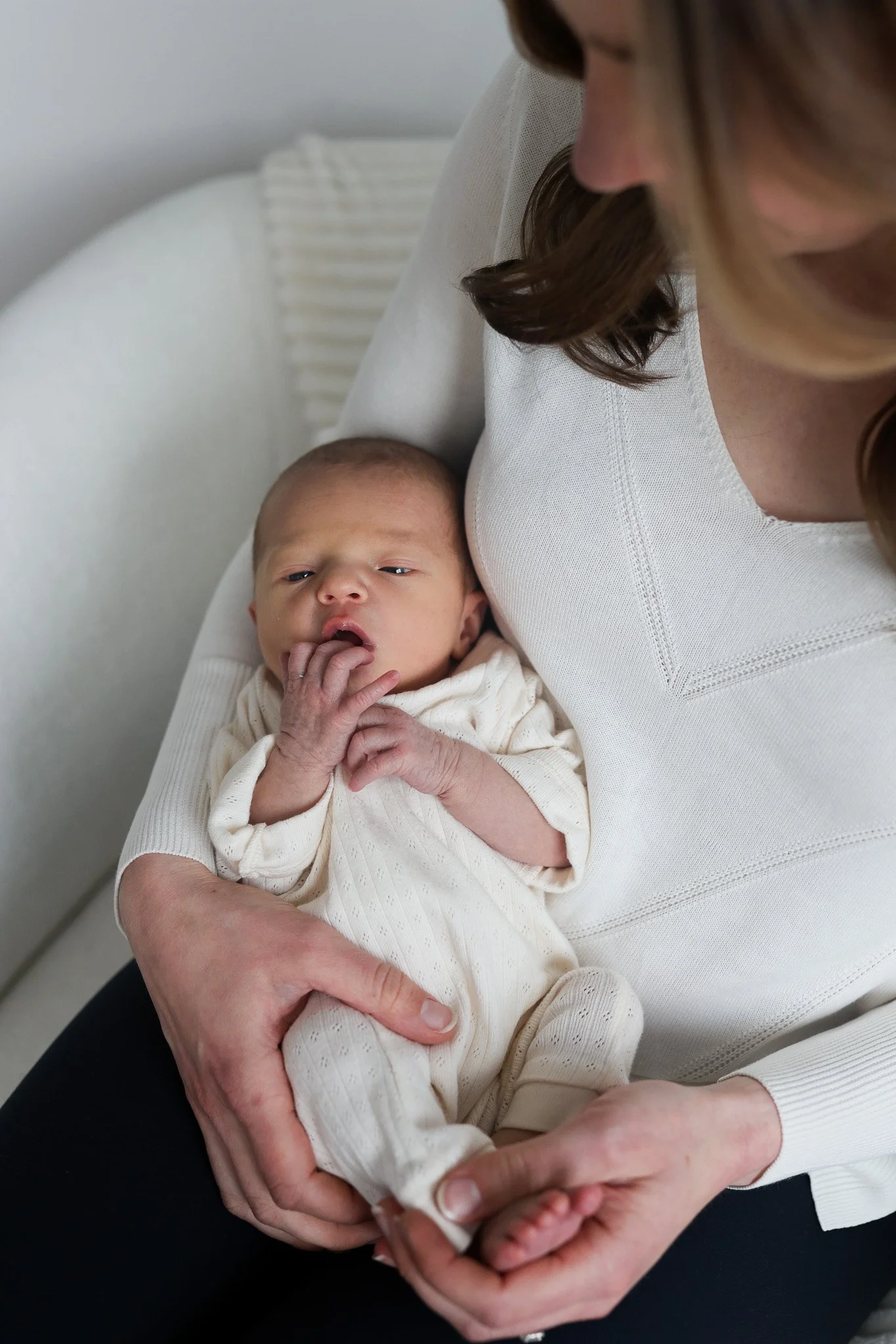 A woman holding a newborn baby in her arms with the baby licking his fingers, white background, woman wearing a white top, baby dressed in light-colored clothing.